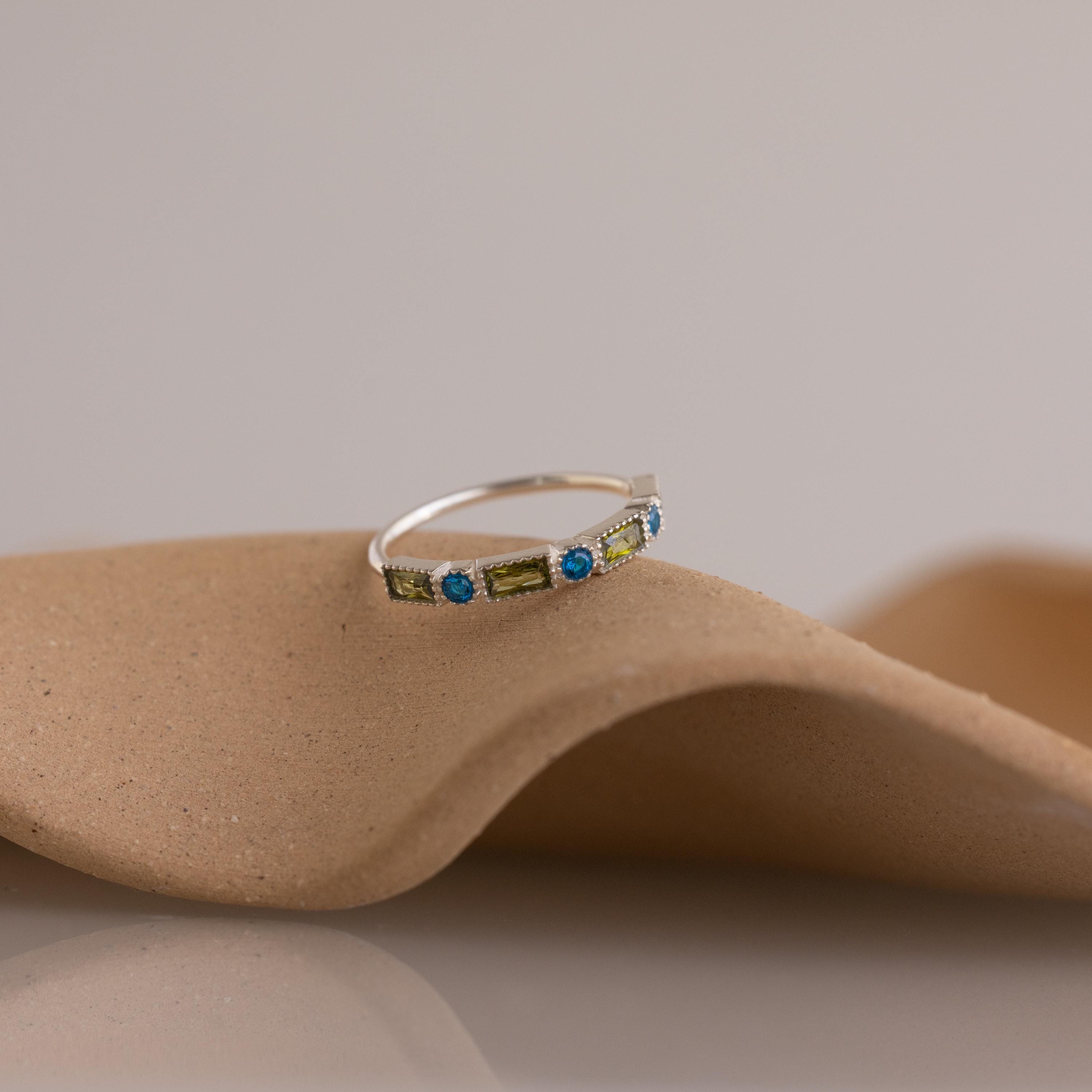 The Mixed Art Deco Birthstone Ring in Sterling Silver features green and blue stones, displayed on a curved beige surface—an ideal choice for anyone seeking a unique personalized birthstone ring.