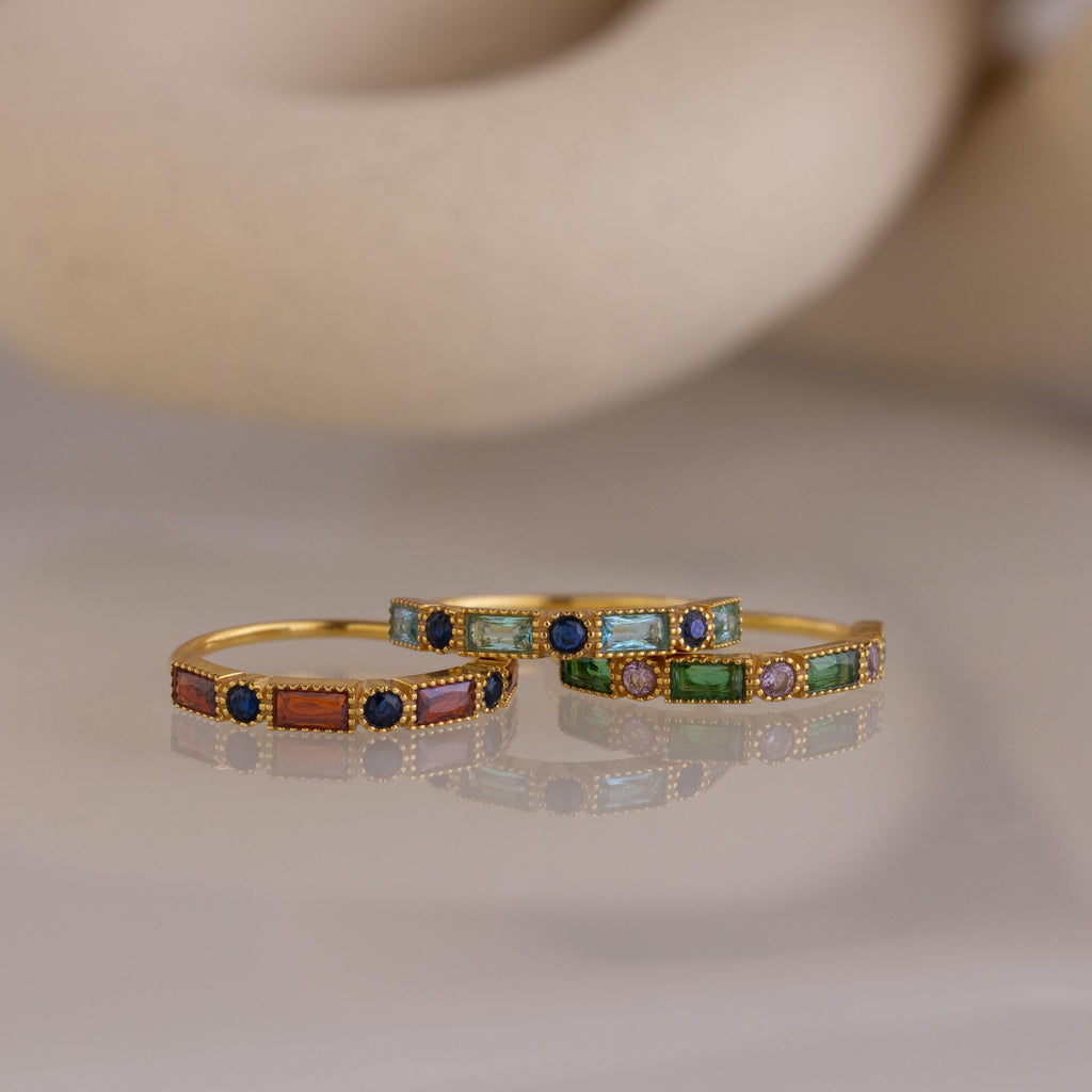 Three Mixed Art Deco Birthstone Rings, featuring colorful rectangular gemstones and small round blue stones, are elegantly displayed on a smooth surface.
