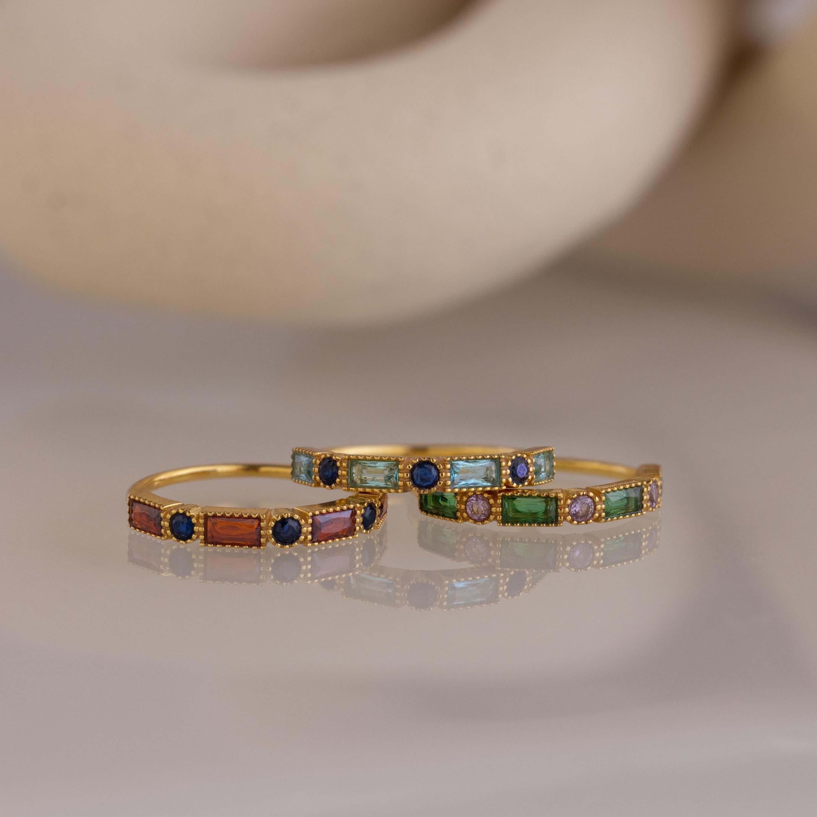 Three Mixed Art Deco Birthstone Rings, featuring colorful rectangular gemstones and small round blue stones, are elegantly displayed on a smooth surface.