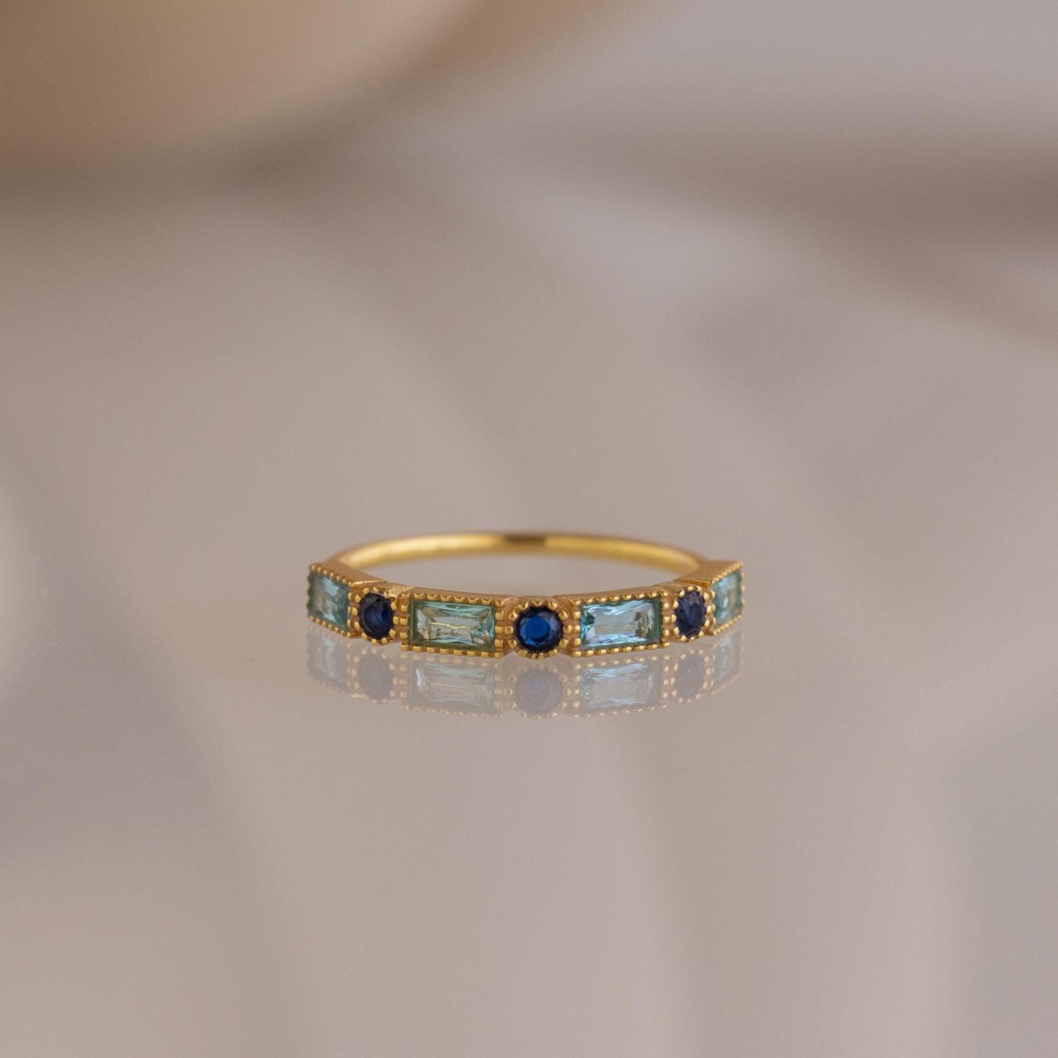 The Mixed Art Deco Birthstone Ring features alternating rectangular aqua and round blue stones in a gold setting, resting on a reflective surface—ideal for fans of dainty birthstone jewelry.