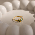 The Toi et Moi Heart Birthstone Ring in 18K Gold, featuring two green gemstones, is displayed in sunlight inside a white scalloped ceramic dish.