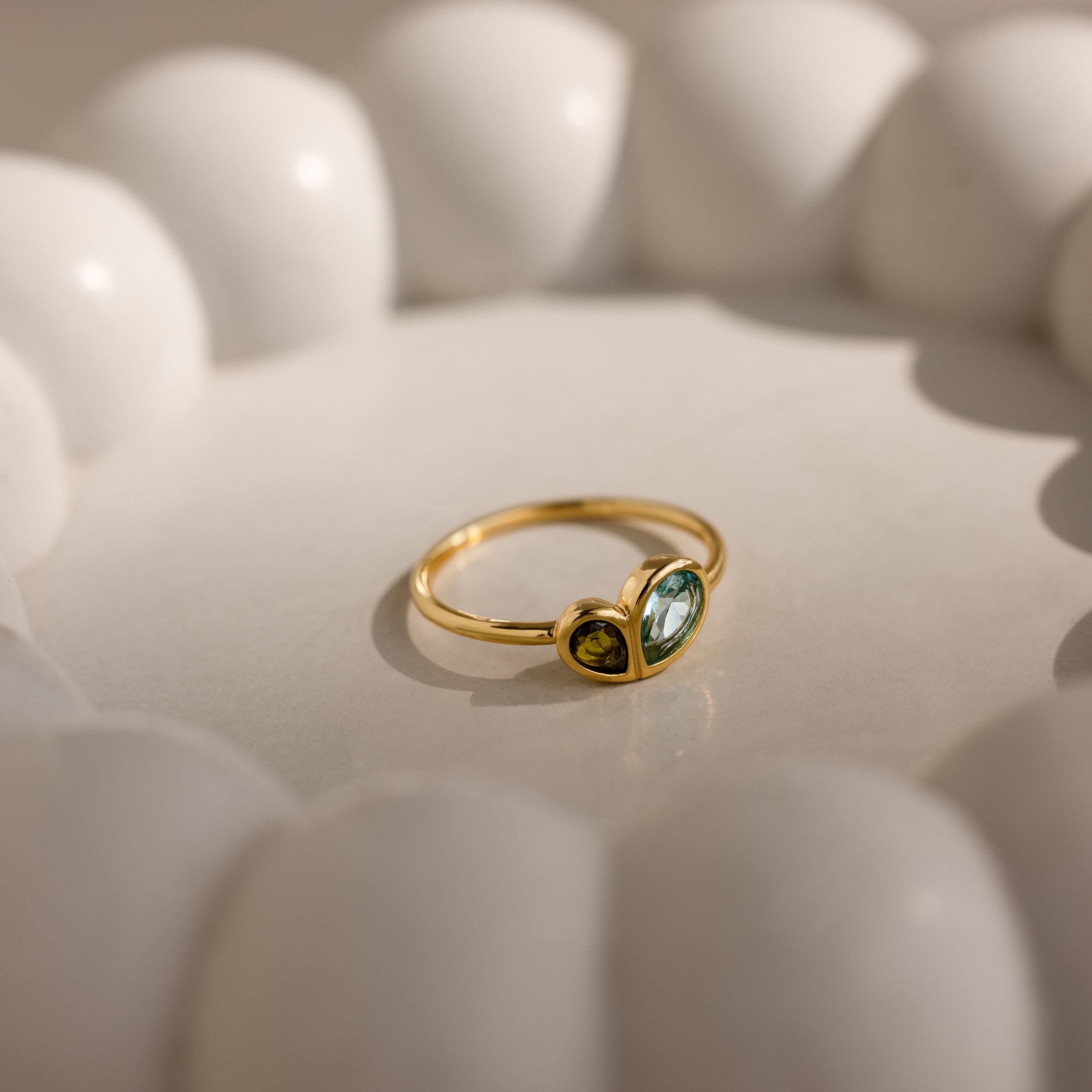 The Toi et Moi Heart Birthstone Ring in 18K Gold, featuring two green gemstones, is displayed in sunlight inside a white scalloped ceramic dish.