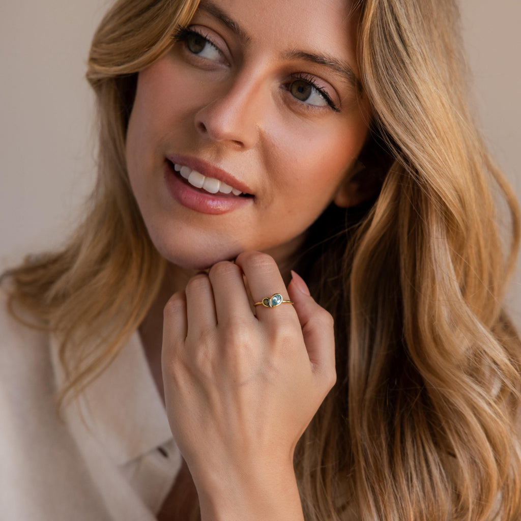 A woman with long blonde hair smiles gently, showing off the Toi et Moi Heart Birthstone Ring in 18K Gold with a blue stone on her hand near her face.