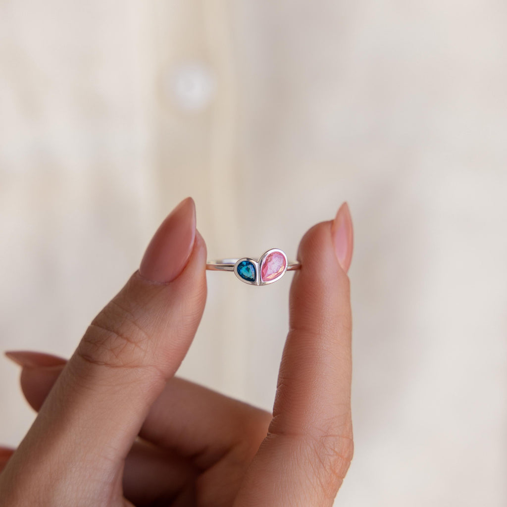 A hand holds the Toi et Moi Heart Birthstone Ring in Sterling Silver, featuring two heart-shaped stones—one blue and one pink.