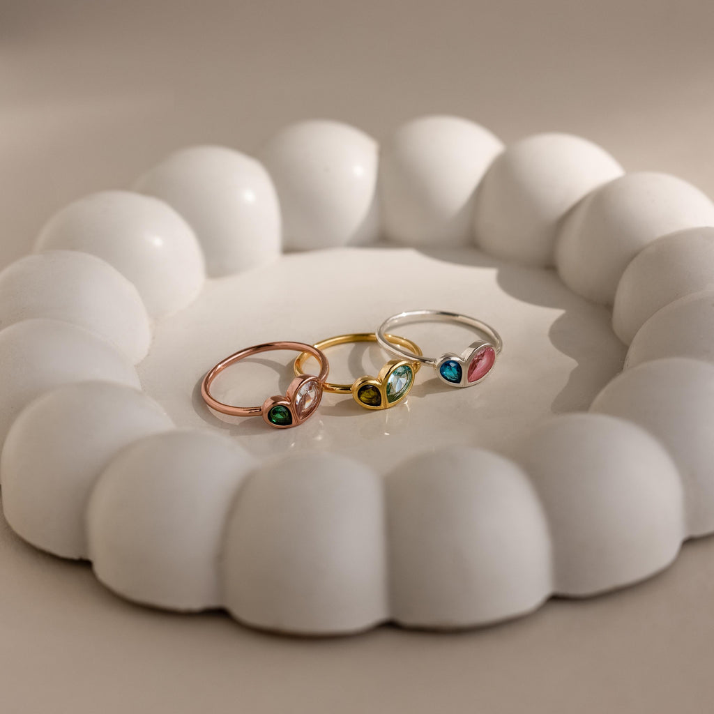 The Toi et Moi Heart Birthstone Ring in Rose Gold, along with two other colorful gemstone rings, is displayed on a round white scalloped ceramic dish.