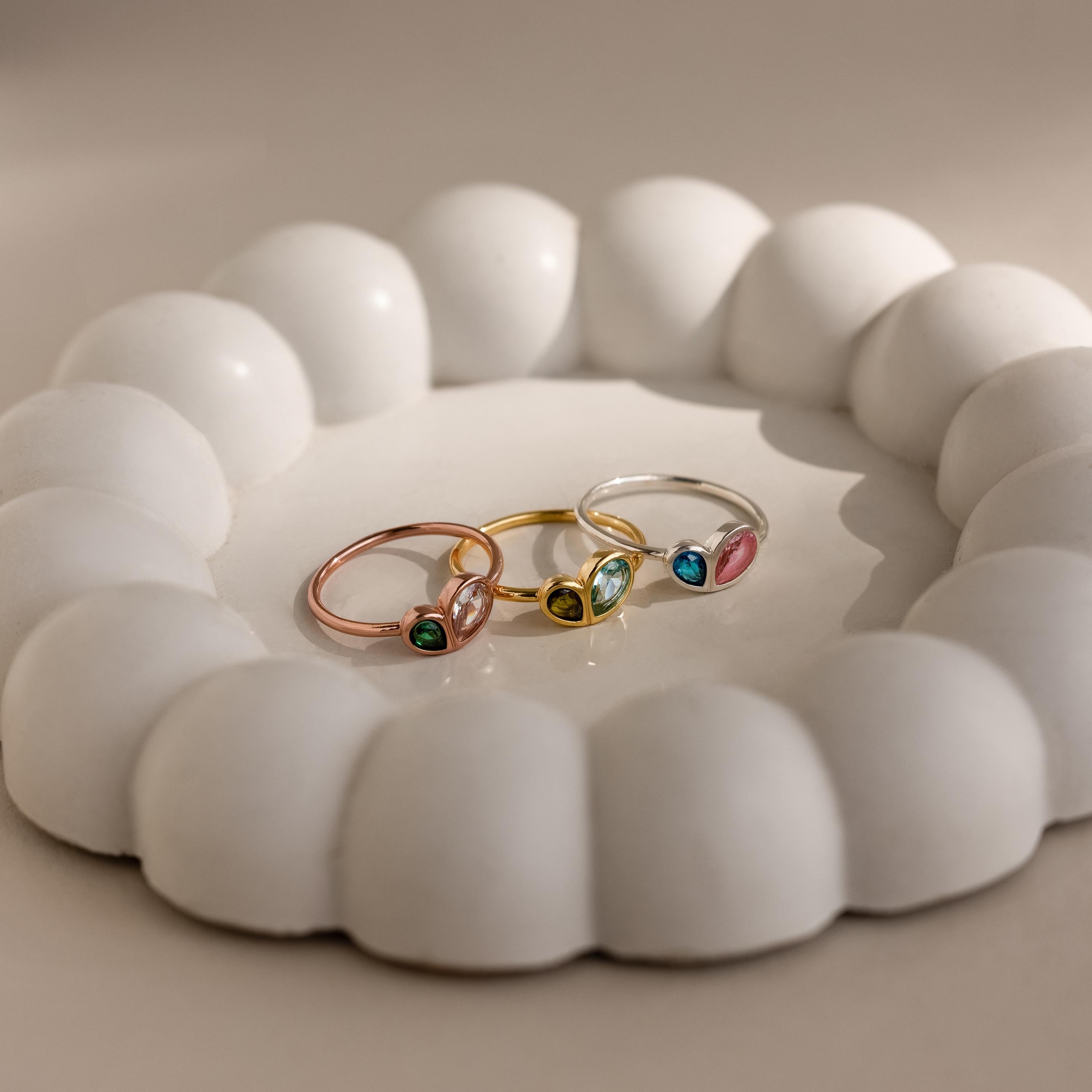 The Toi et Moi Heart Birthstone Ring in Rose Gold, along with two other colorful gemstone rings, is displayed on a round white scalloped ceramic dish.