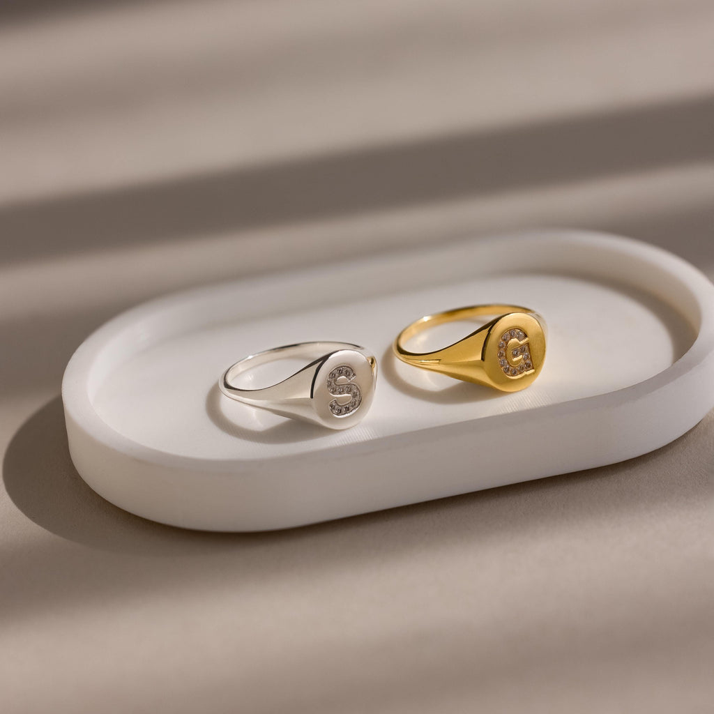 The Pave Initial Signet Ring, available in silver or gold with an engraved letter S, rests on a white oval tray—an ideal statement piece to personalize your style.