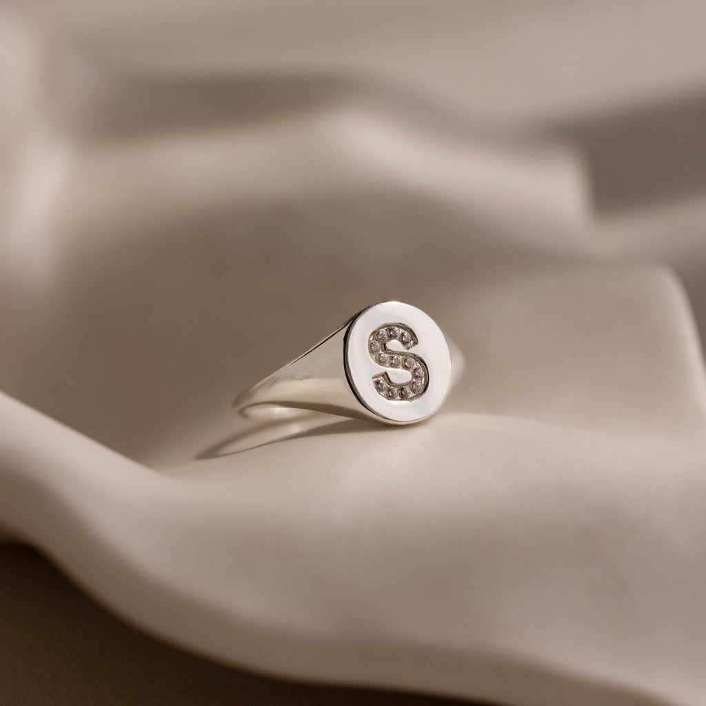 The Pave Initial Signet Ring in Sterling Silver, featuring a crystal-studded "S" on a round face, is displayed elegantly on soft, cream fabric.