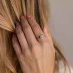 A hand with light pink nails wears the Toi et Moi Heart Birthstone Ring, a gold promise ring featuring a blue pear-shaped gemstone and a small brown accent.