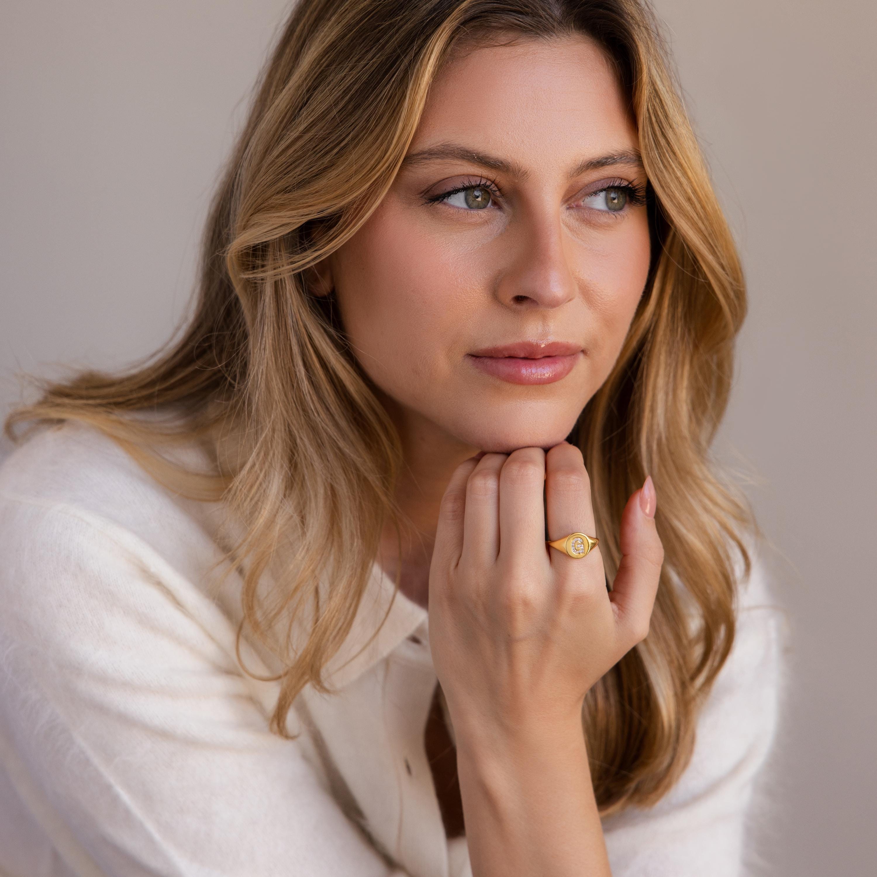 Woman with wavy blonde hair, wearing a white top and the Pave Initial Signet Ring, gazes thoughtfully to the side.
