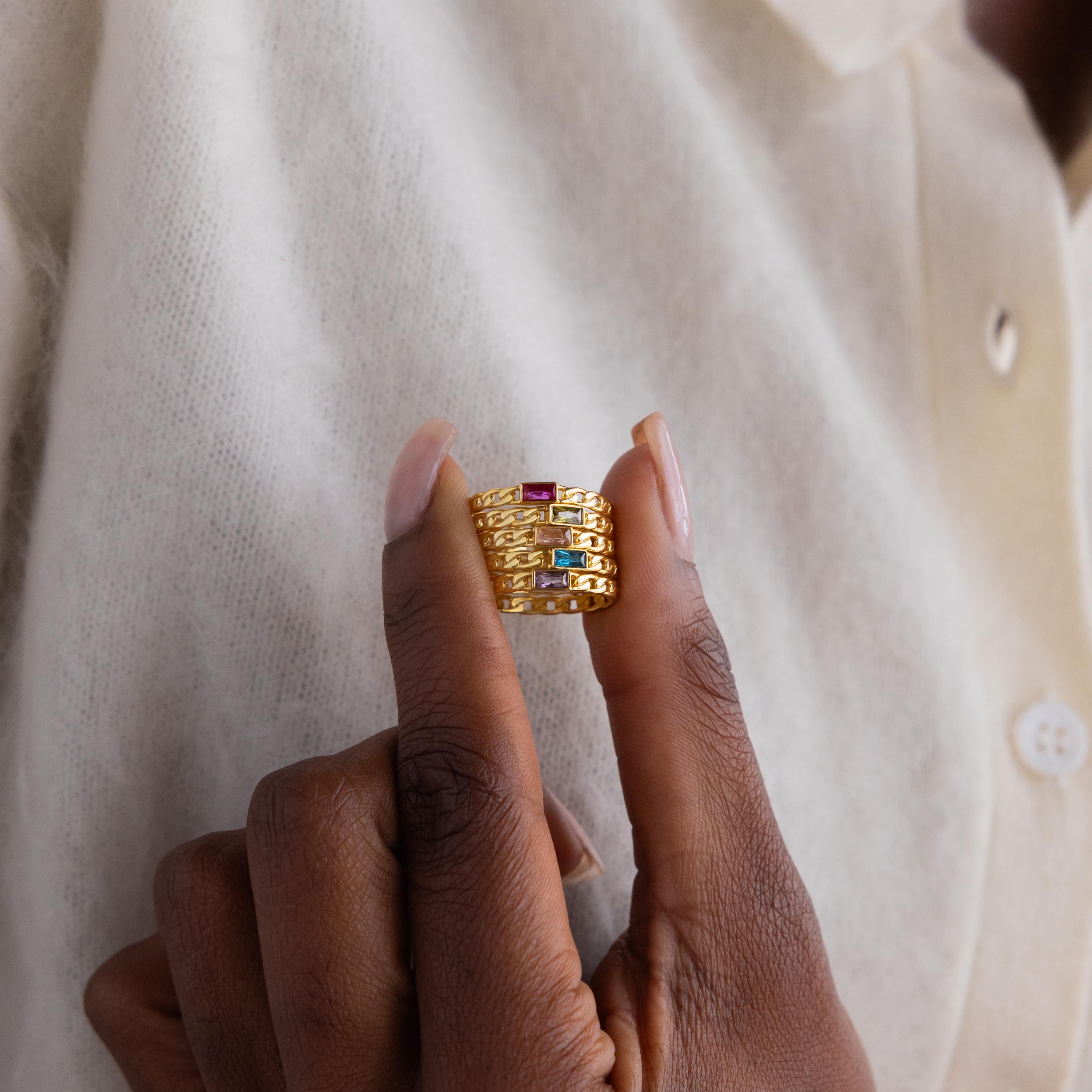 A hand displays the Baguette Topaz Curb Ring, featuring bezel-set gemstones, against a cream shirt background.