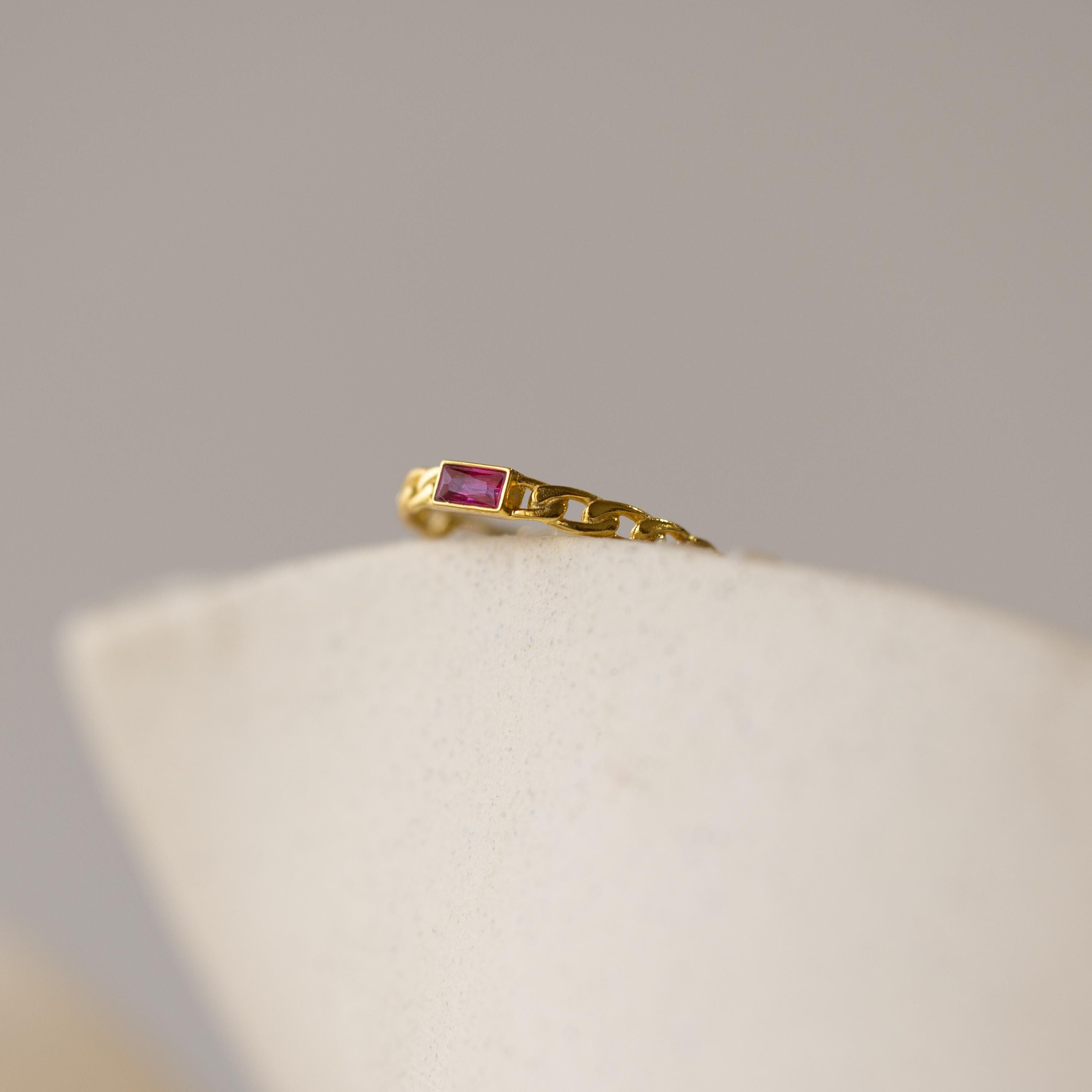 The Baguette Topaz Curb Ring features a gold chain band with a bezel-set rectangular red stone, displayed on a white curved surface.