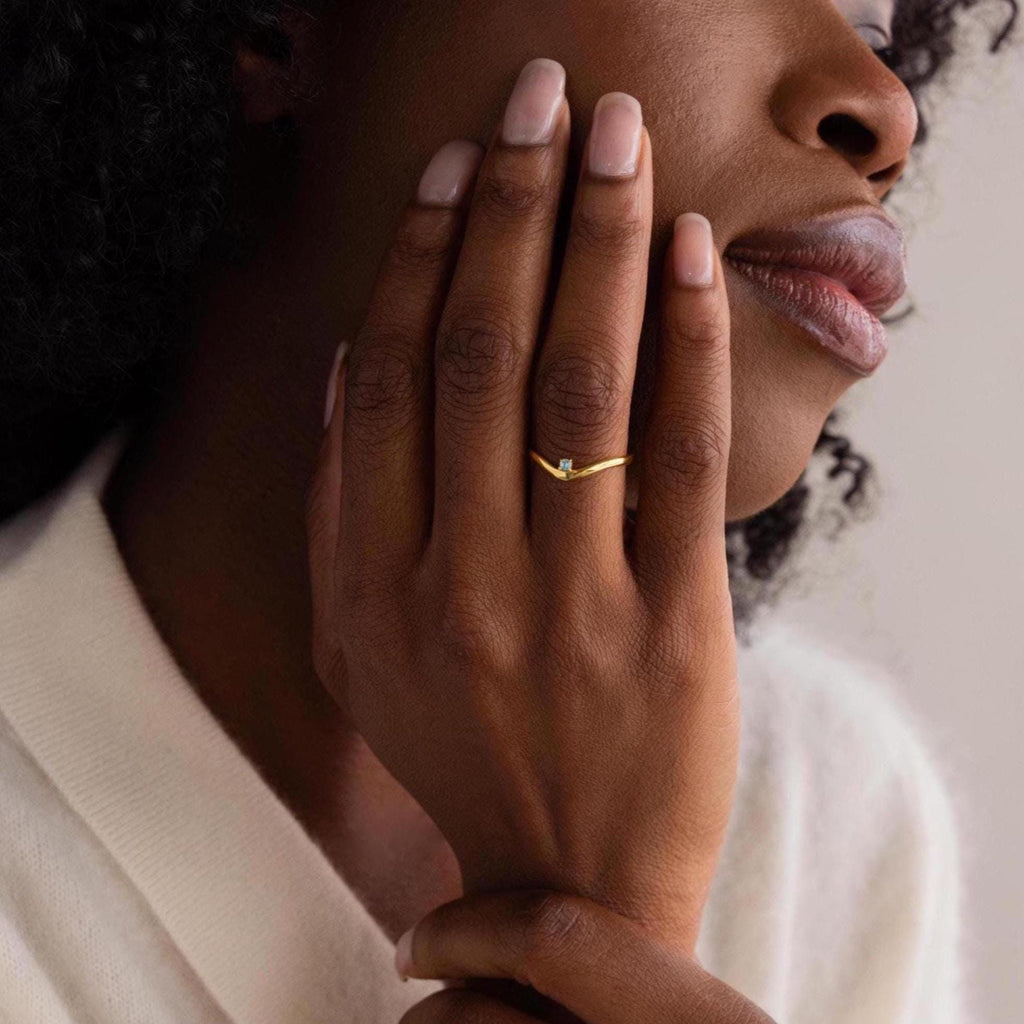 A woman touches her face, showcasing the Onyx Drop Curve Ring—a delicate gold band with a small onyx gemstone adorning her finger.