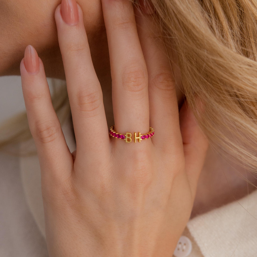 A hand with manicured nails wears the Duo Initial Onyx Tennis Ring, a personalized eternity band featuring pink beads and gold letters B and H.