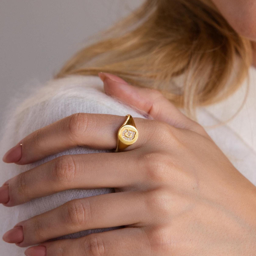 A woman showcases the elegance of the Pave Initial Signet Ring in 18K Gold, featuring a decorative engraving on her index finger.