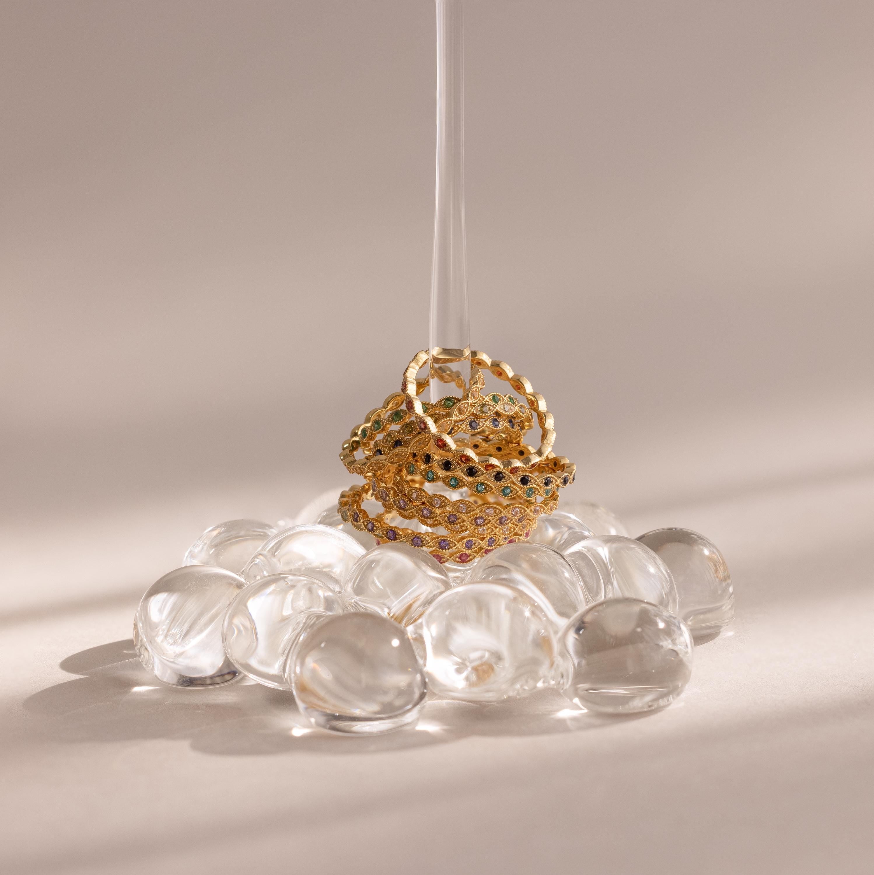 The Dainty Marquise Topaz Ring shines atop gold gemstone bands and clear glass pebbles, pierced by a vertical glass rod for an Art Deco-inspired display.