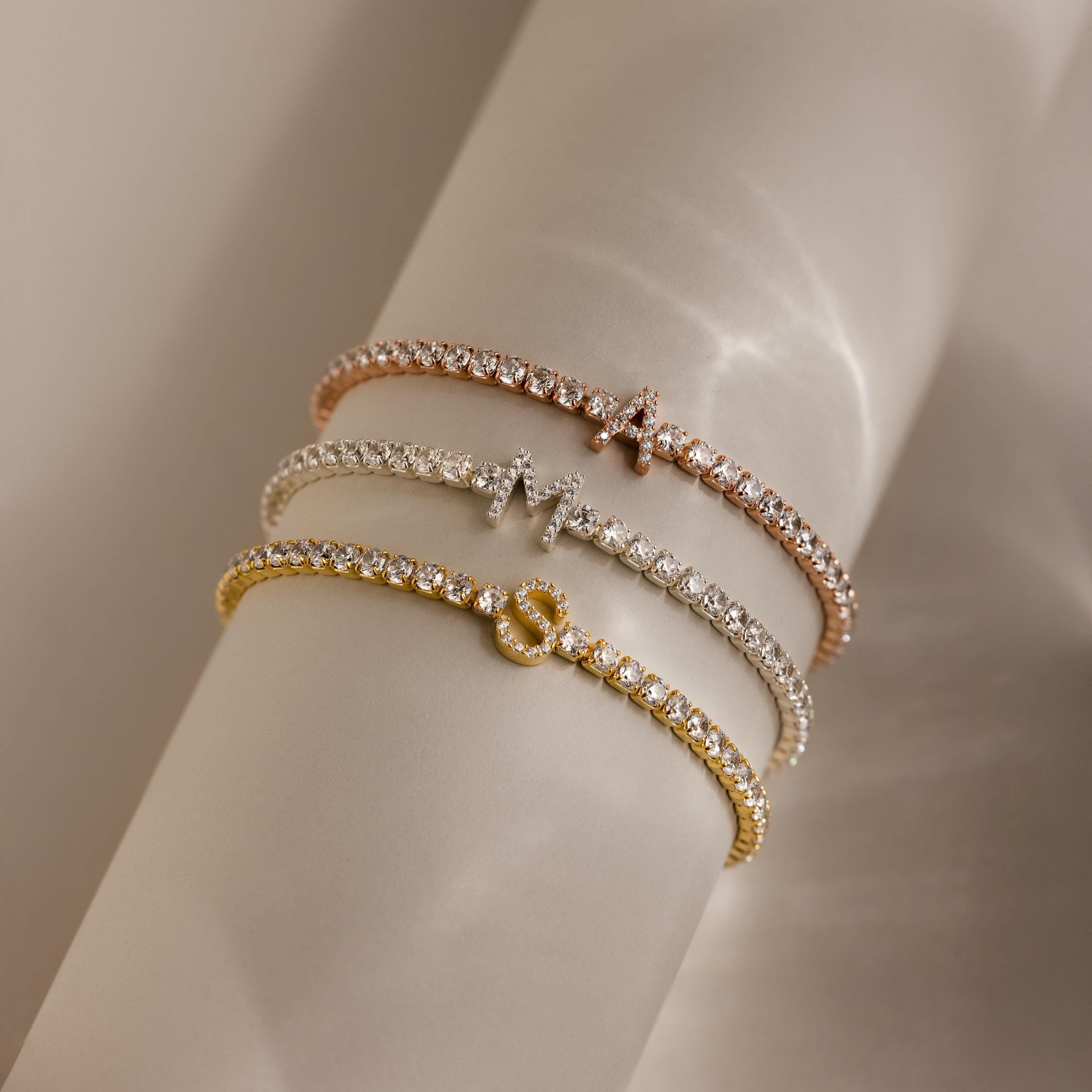 Three Diamond Tennis Initial Bracelets, each with a custom initial charm—A, M, and S—sparkle on a softly lit beige background.