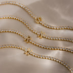 Four stackable bracelets, including the Diamond Tennis Initial Bracelet, are displayed on a beige surface with soft shadows. Personalize your look by adding your custom initial to the sparkling design.