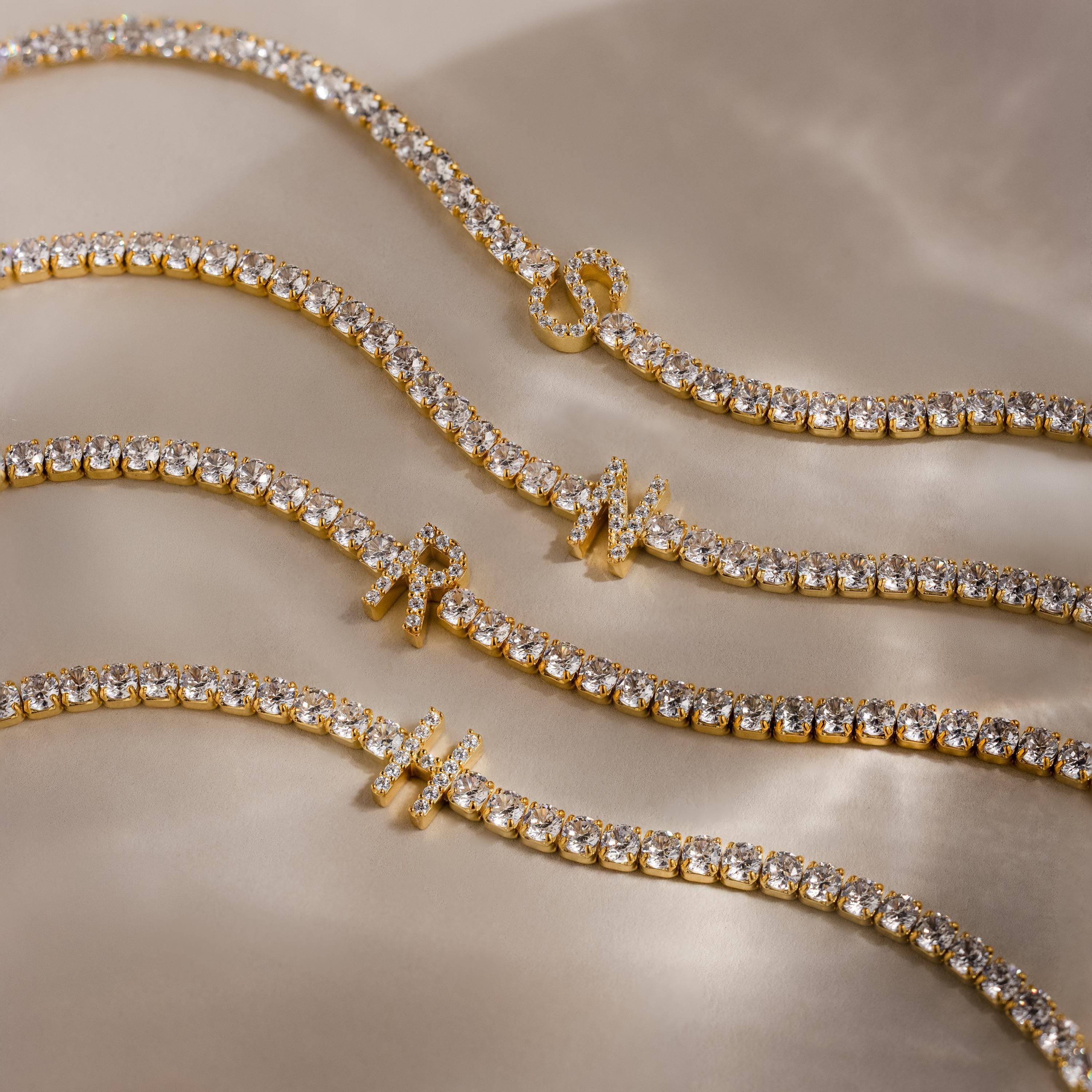 Four stackable bracelets, including the Diamond Tennis Initial Bracelet, are displayed on a beige surface with soft shadows. Personalize your look by adding your custom initial to the sparkling design.