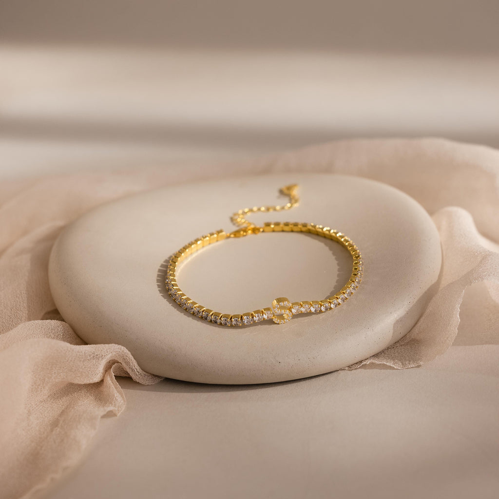 The Diamond Tennis Initial Bracelet, featuring clear stones, is shown on a smooth oval stone with a soft fabric backdrop—this stackable piece adds personalized elegance to your jewelry collection.
