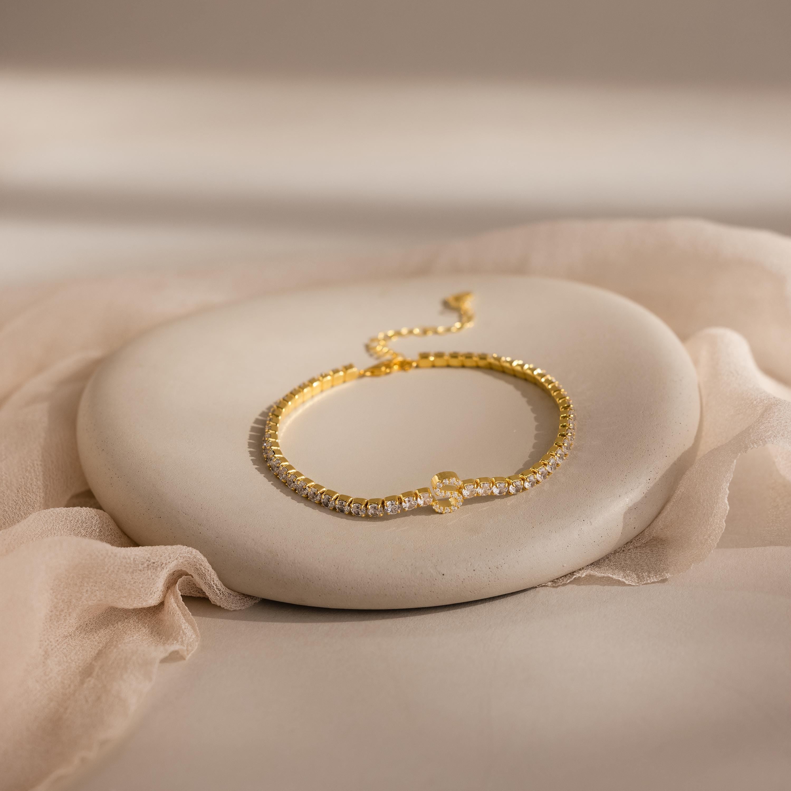 The Diamond Tennis Initial Bracelet, featuring clear stones, is shown on a smooth oval stone with a soft fabric backdrop—this stackable piece adds personalized elegance to your jewelry collection.