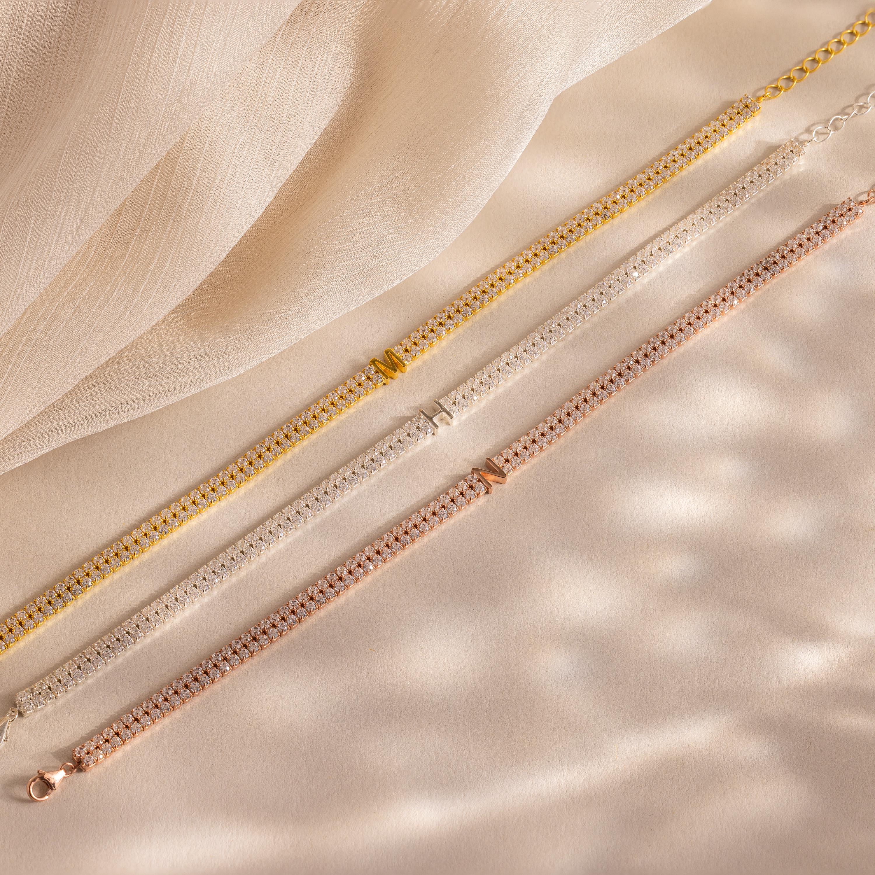 The Initial Duo Tennis Bracelet, featuring gold, silver, and rose gold chains, is elegantly displayed on soft beige fabric—ideal for layering or customizing with your chosen initial for a personal touch.