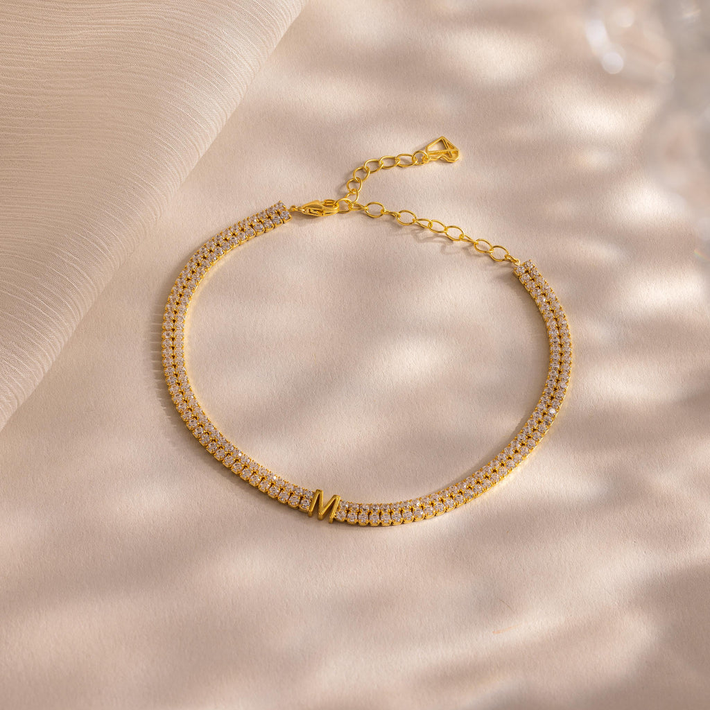 Gold necklace with small diamonds and a custom initial M charm on a soft beige surface. Complete your look by pairing it with our Initial Duo Tennis Bracelet for an elegant, personalized style.