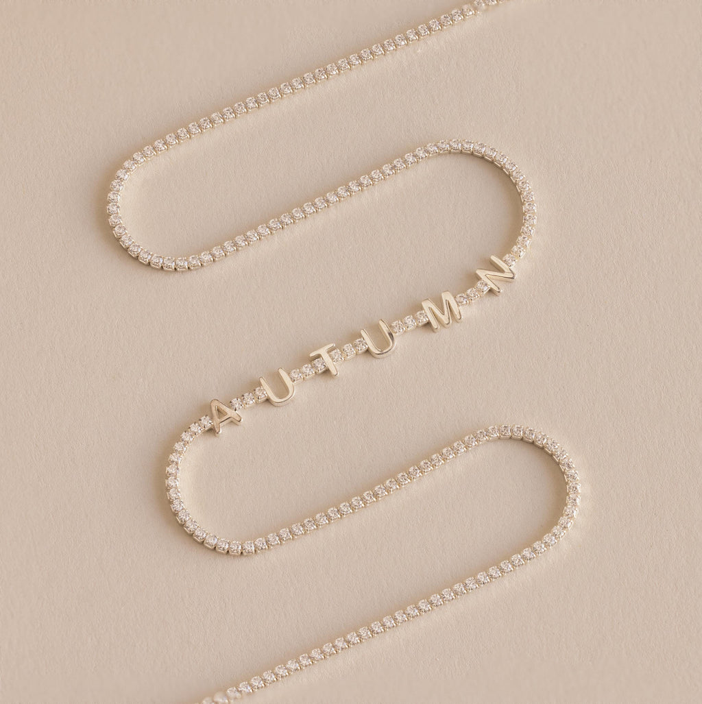 The Diamond Tennis Letter Necklace features gold letter charms spelling AUTUMN with small diamonds, styled on a beige background. This elegant piece is inspired by the classic tennis necklace design.