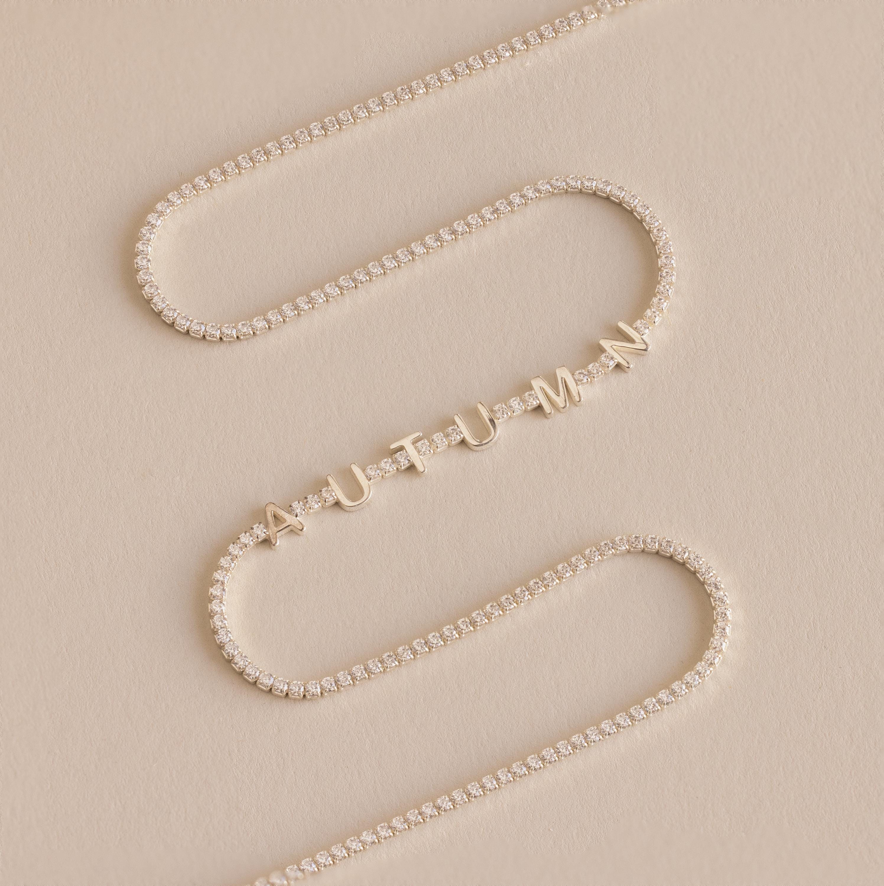 The Diamond Tennis Letter Necklace features gold letter charms spelling AUTUMN with small diamonds, styled on a beige background. This elegant piece is inspired by the classic tennis necklace design.