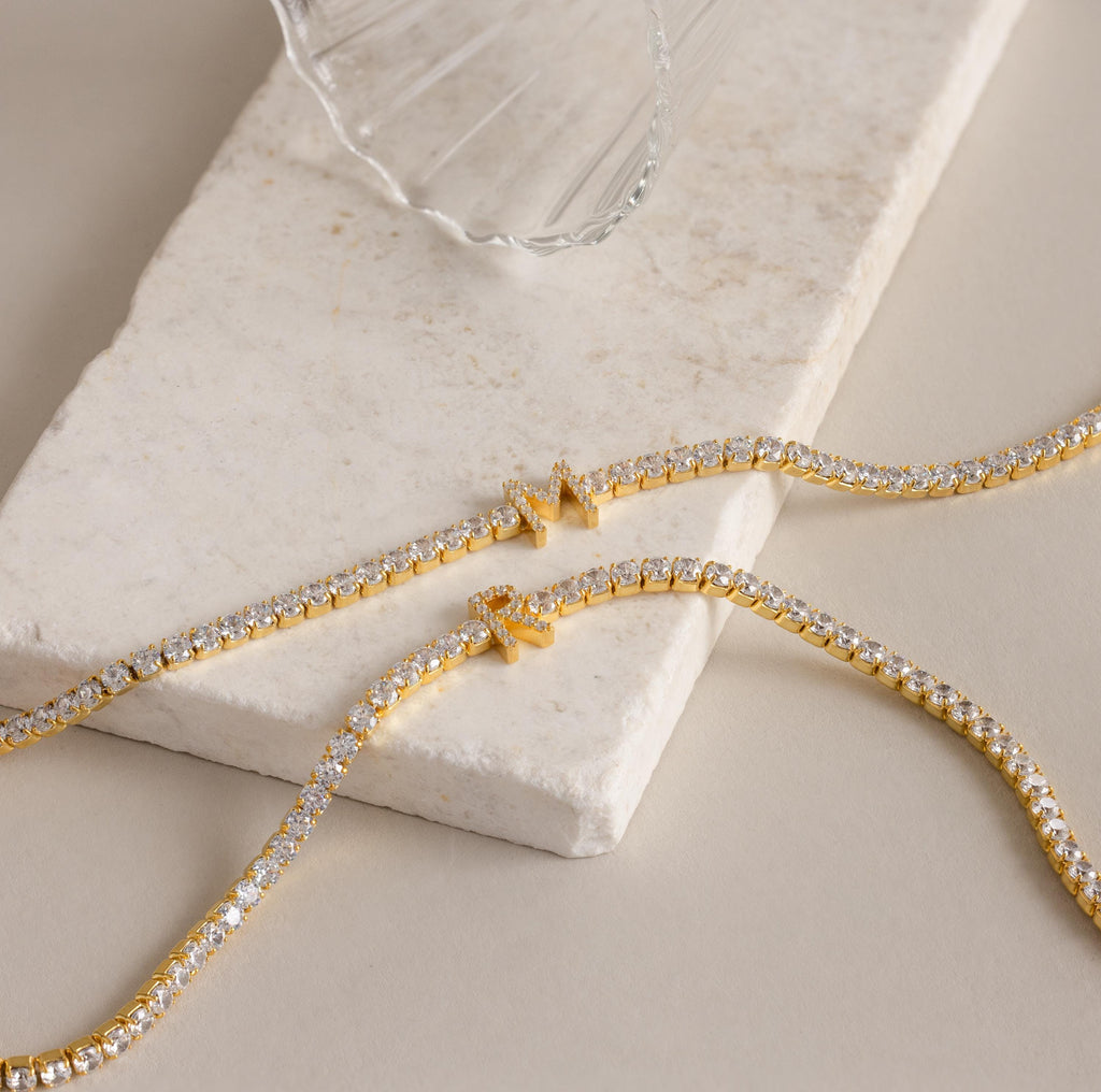 Two Diamond Tennis Initial Necklaces—ideal as personalized jewelry—are displayed on a marble slab next to a clear glass.
