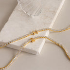 Two Diamond Tennis Initial Necklaces—ideal as personalized jewelry—are displayed on a marble slab next to a clear glass.