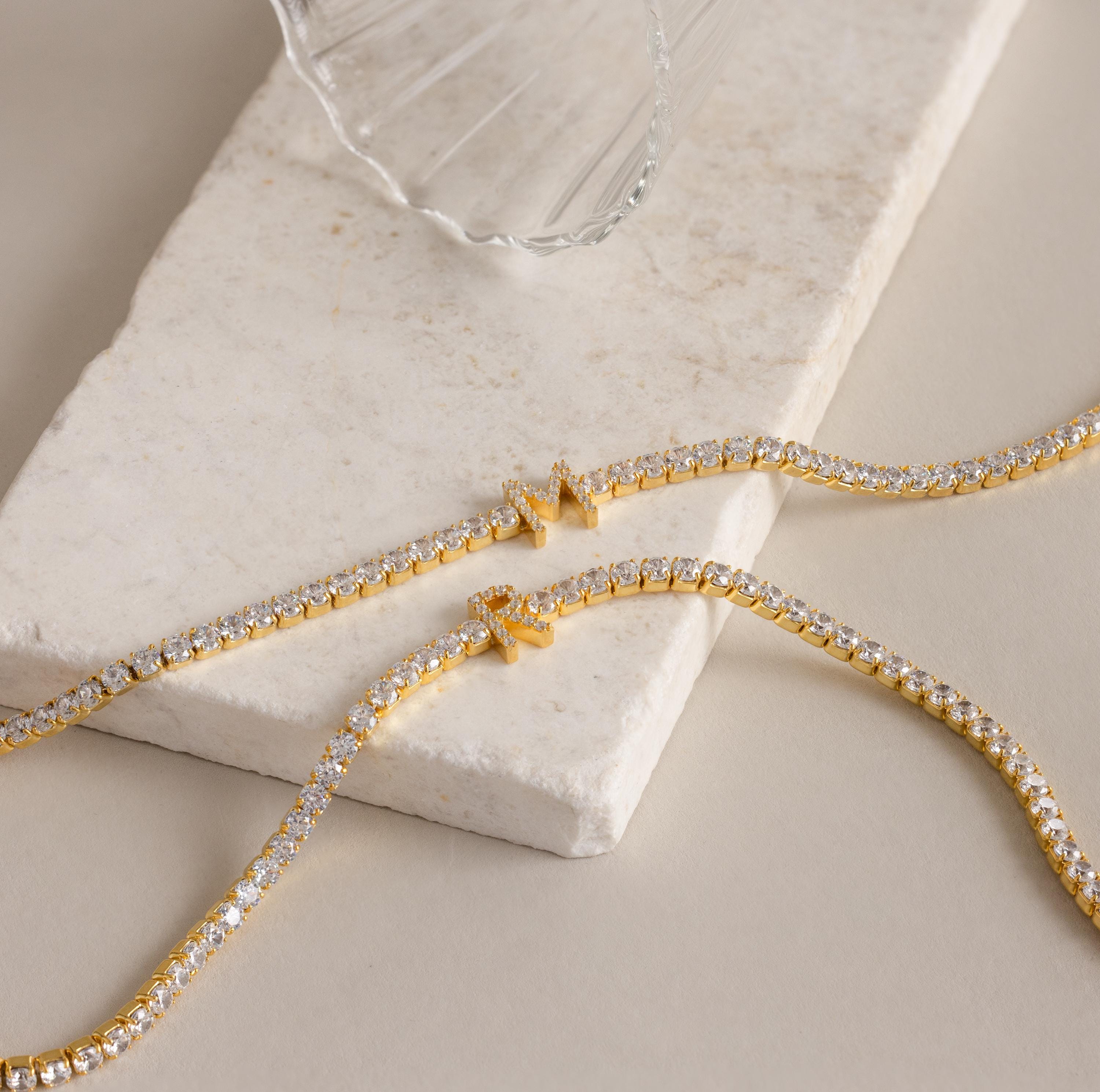 Two Diamond Tennis Initial Necklaces—ideal as personalized jewelry—are displayed on a marble slab next to a clear glass.