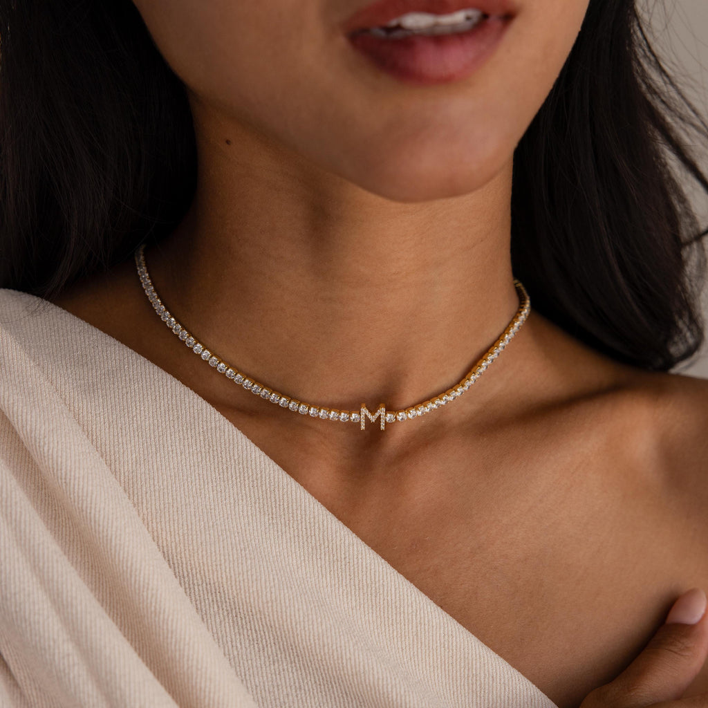A woman in a cream off-shoulder top showcases the elegance of personalized jewelry with the Diamond Tennis Initial Necklace, featuring a gold chain and a single diamond-studded “M” charm.