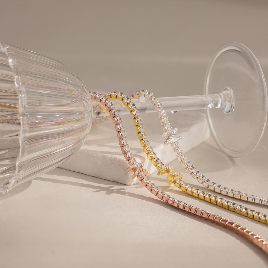 The Diamond Tennis Initial Necklace shines in gold, silver, and rose gold as it drapes elegantly over a tipped wine glass.