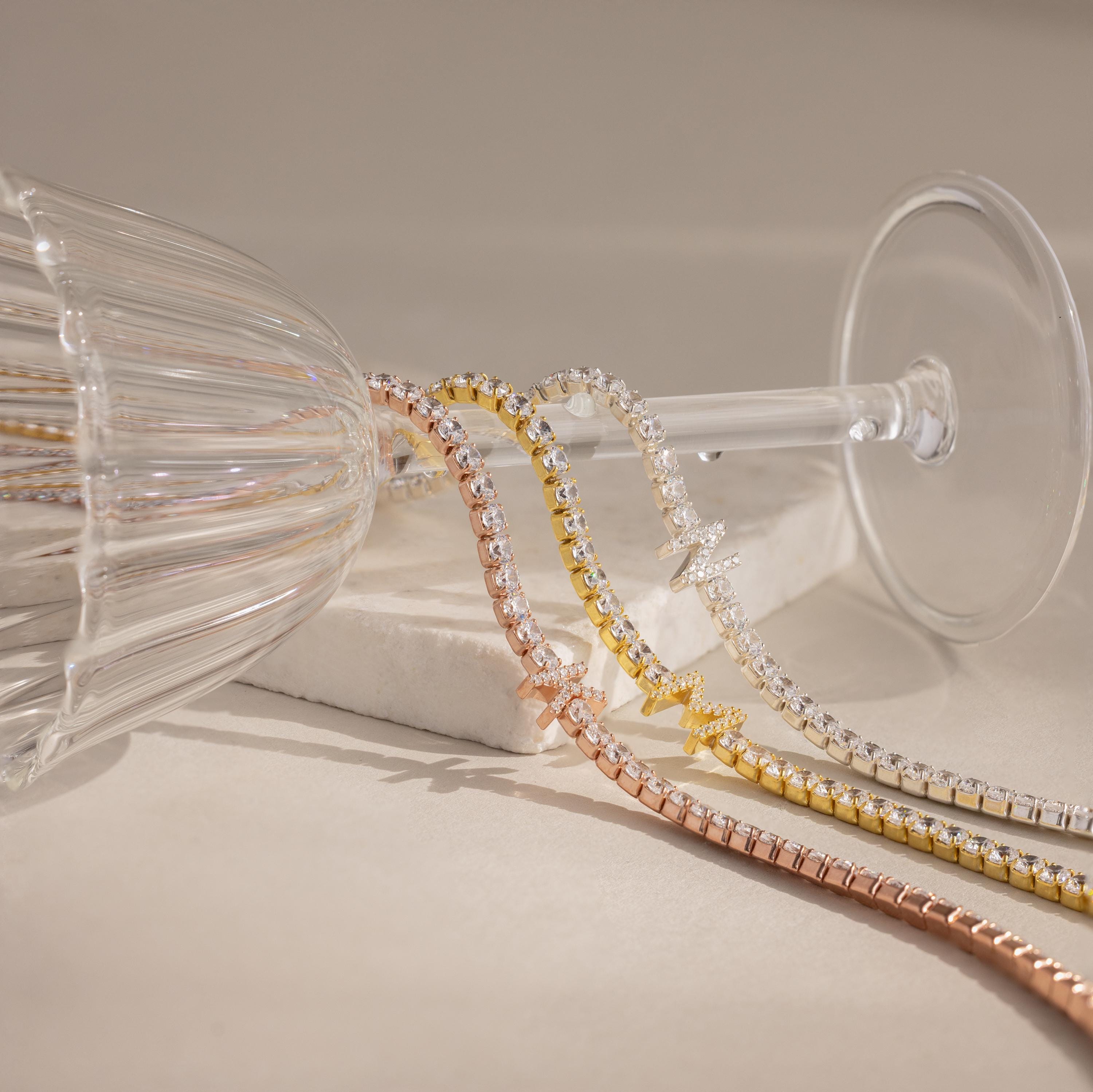 The Diamond Tennis Initial Necklace shines in gold, silver, and rose gold as it drapes elegantly over a tipped wine glass.