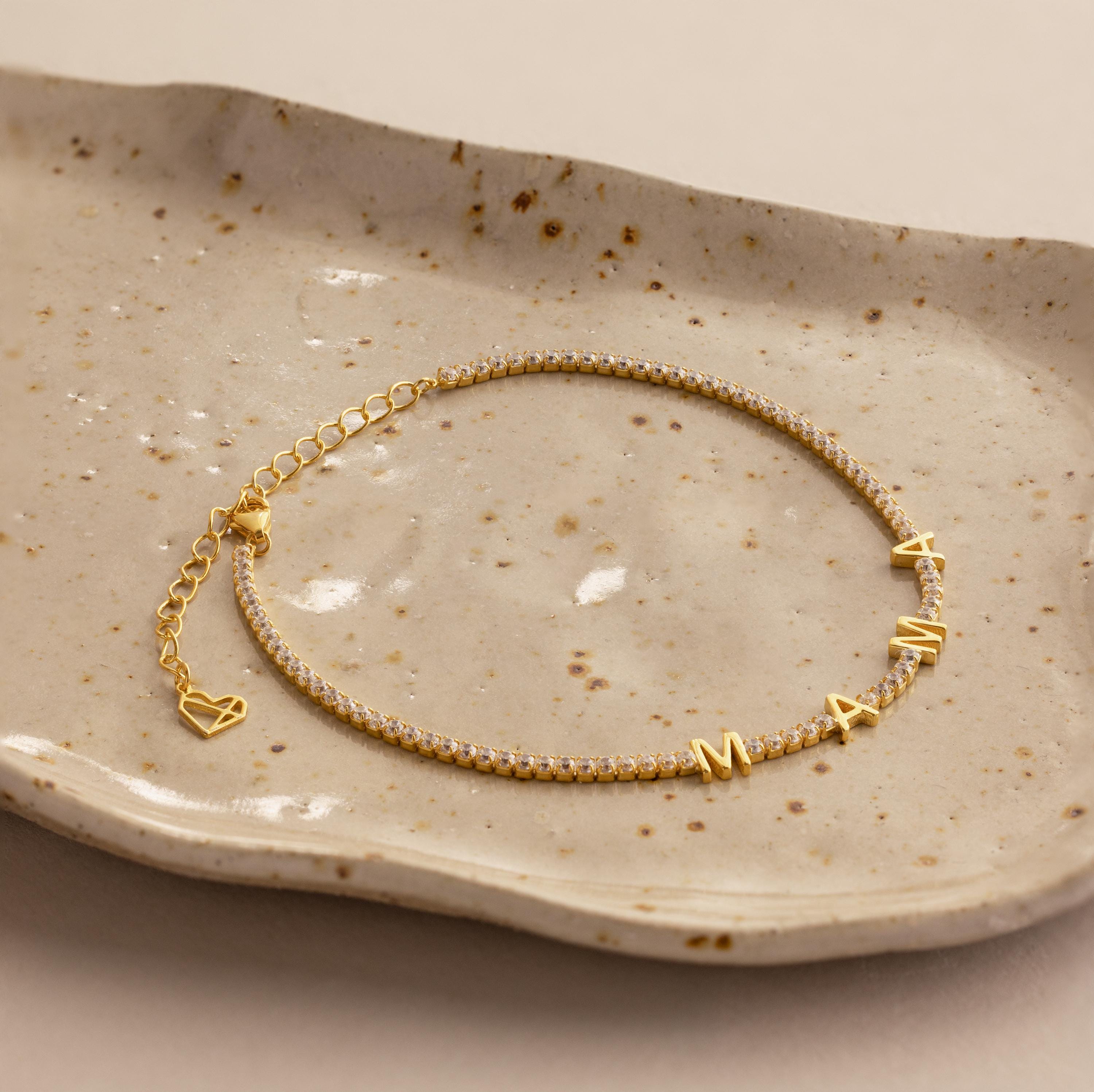 Diamond Tennis Letter Bracelet featuring "MAMA" letter charms and a heart, elegantly displayed on a speckled ceramic dish.