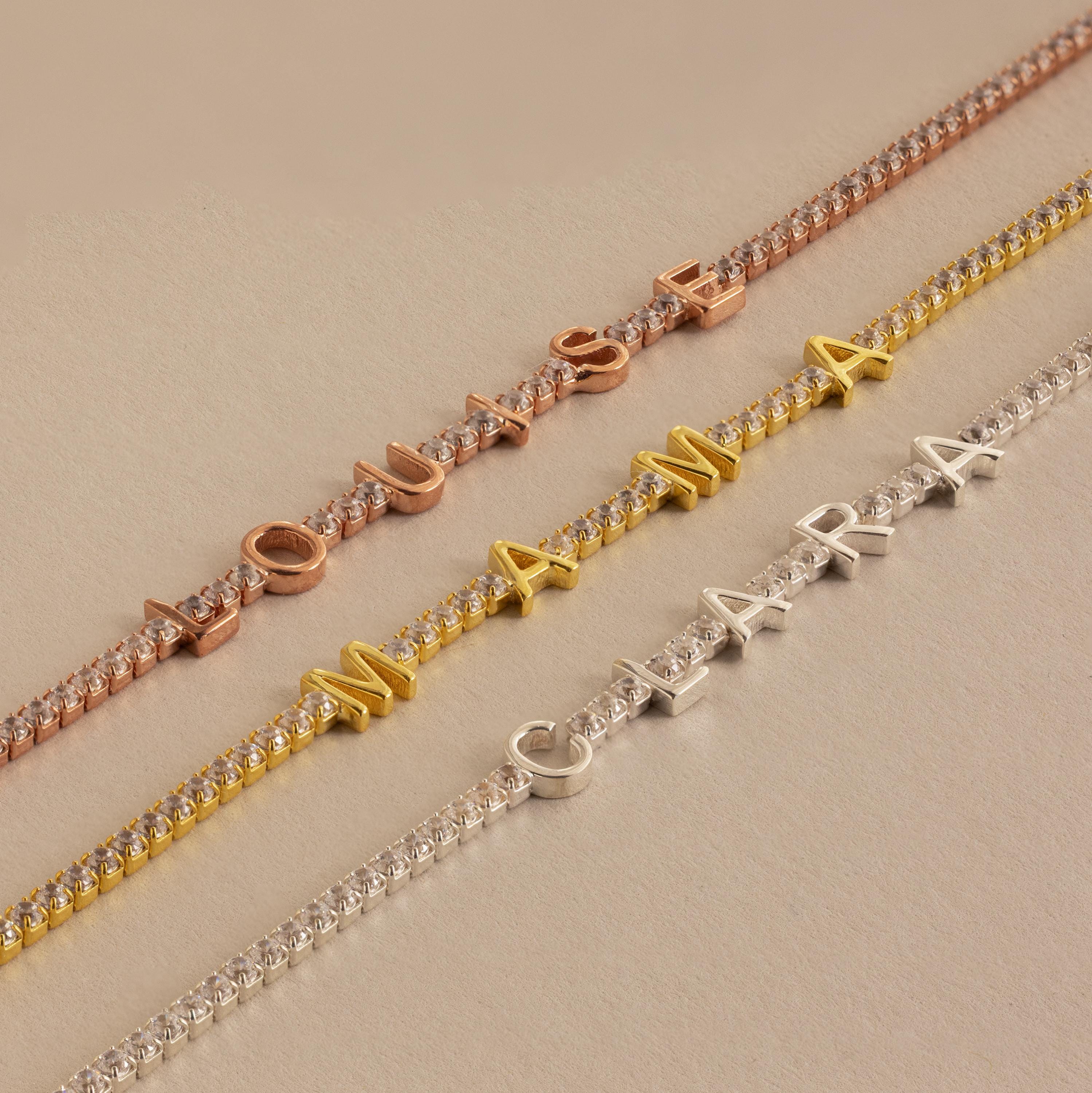 Diamond Tennis Letter Bracelets with names "LOUISE," "MAMA," and "CLARA" in rose, gold, and silver letters rest on a beige background—perfect minimalist personalized accessories.