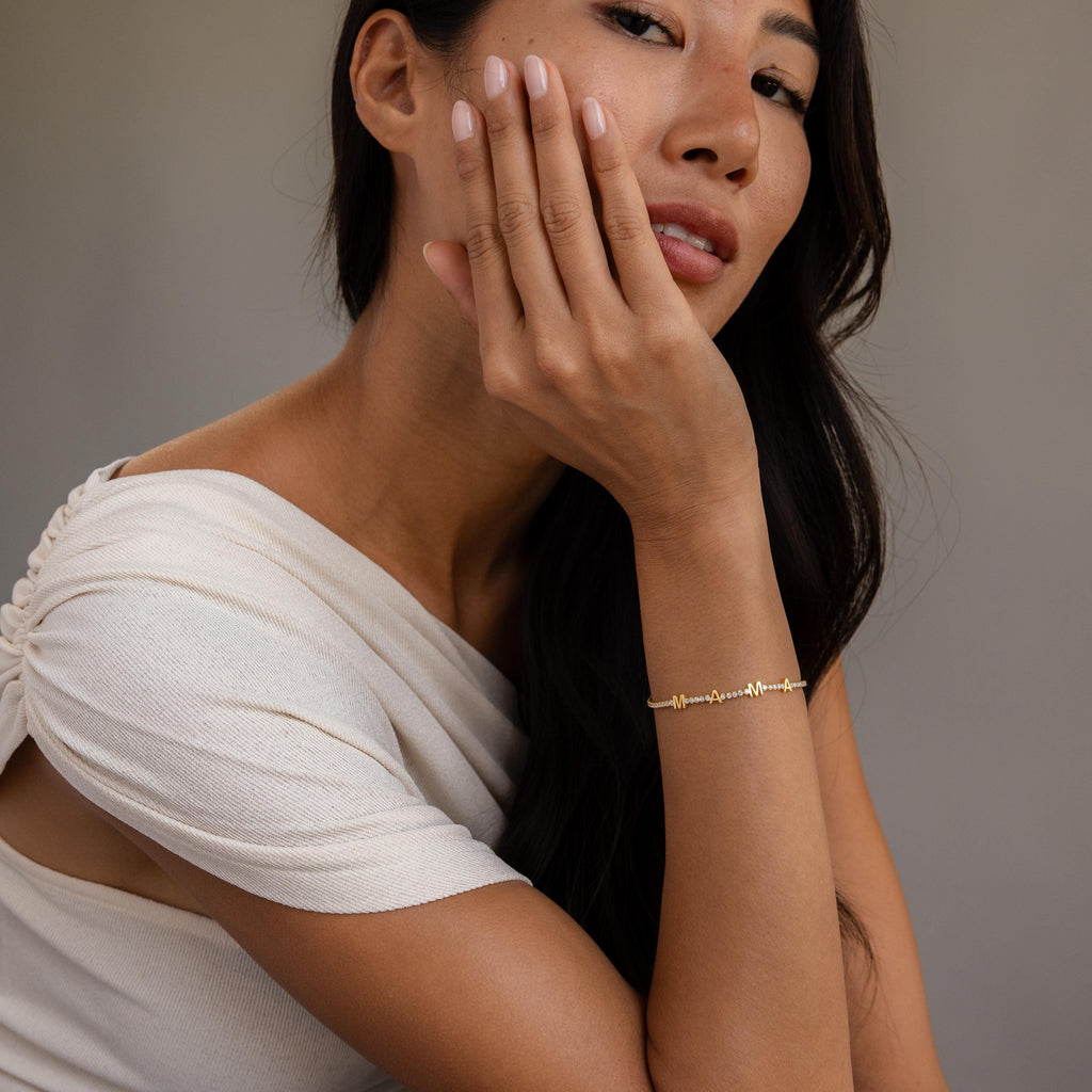 A woman in a white top rests her hand on her face, showcasing the Diamond Tennis Letter Bracelet as she looks at the camera.