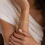 A woman wears a Diamond Tennis Letter Bracelet and a gold ring, paired with a cream off-shoulder top.