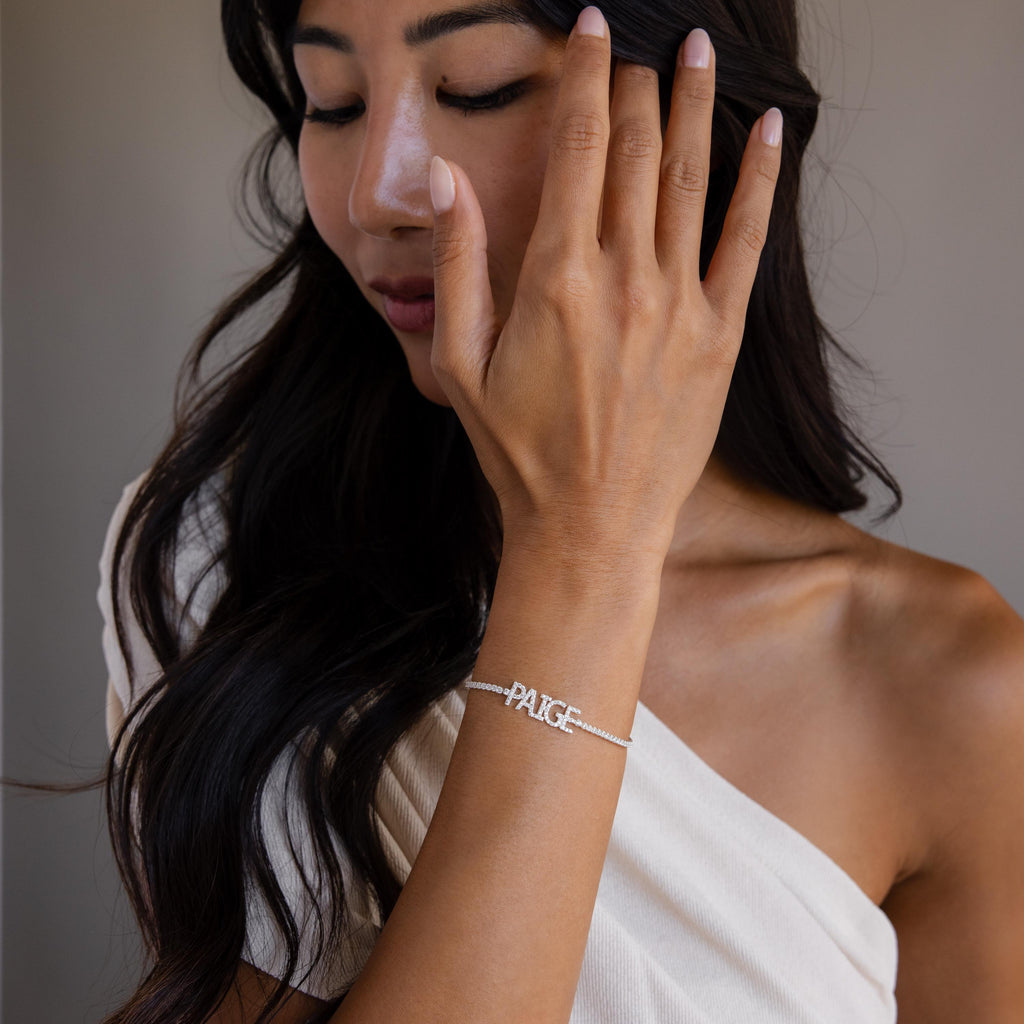 A woman in an off-shoulder top touches her face, showcasing the Pave Name Tennis Bracelet with "PAIGE" on her wrist.