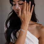 A woman in an off-shoulder top touches her face, showcasing the Pave Name Tennis Bracelet with "PAIGE" on her wrist.