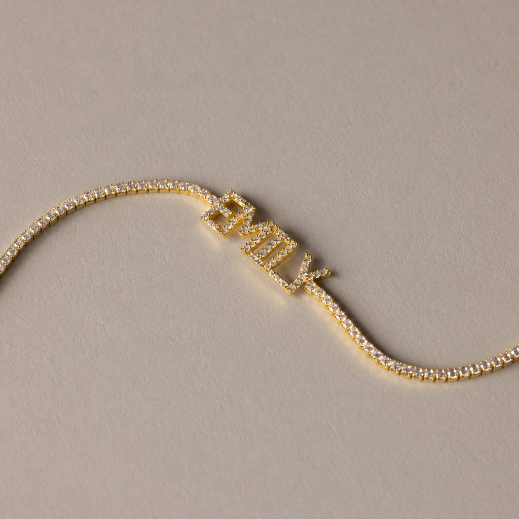 The Pave Name Tennis Bracelet showcases "EMILY" in sparkling diamond-studded letters, creating a stunning personalized bracelet set against a beige background.