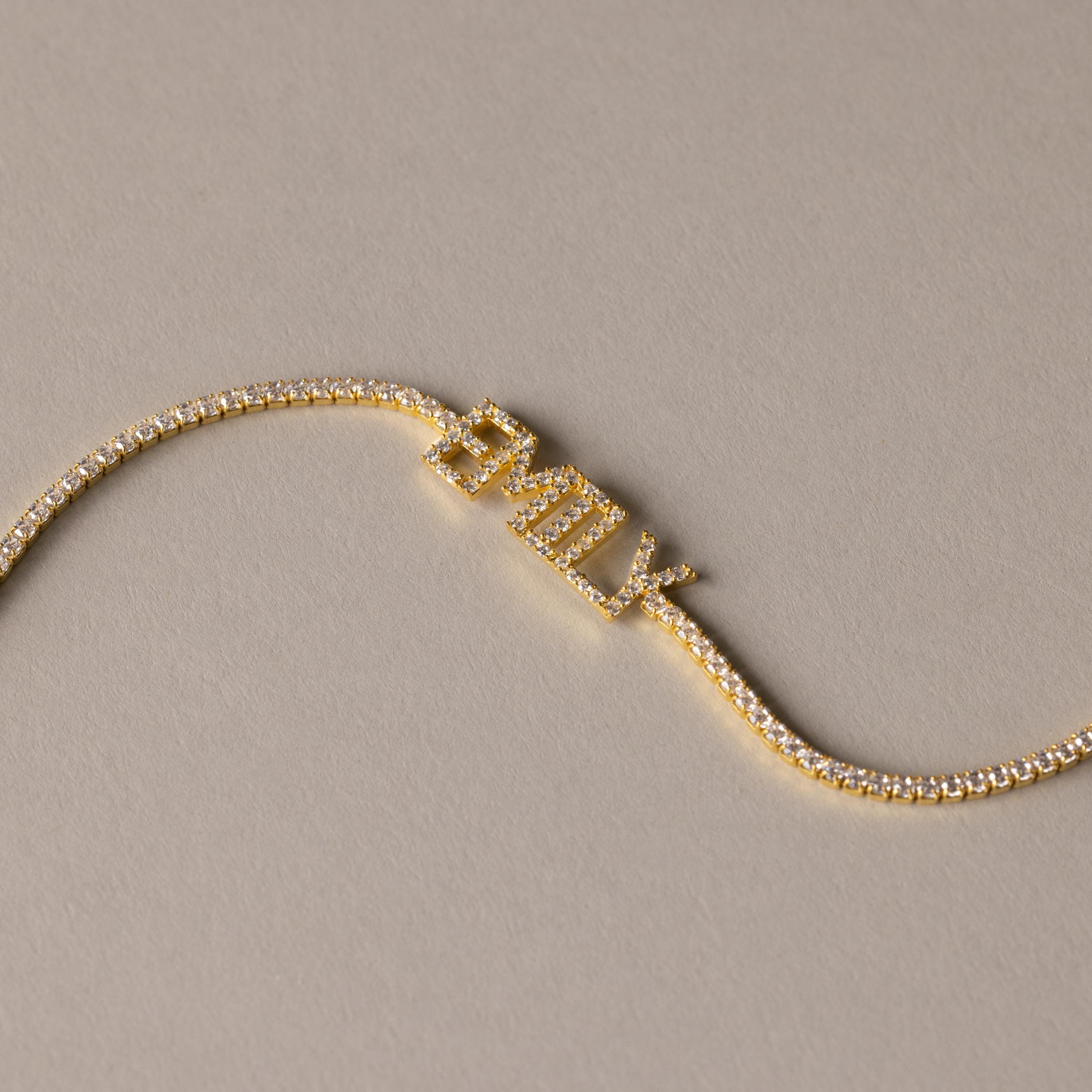 The Pave Name Tennis Bracelet showcases "EMILY" in sparkling diamond-studded letters, creating a stunning personalized bracelet set against a beige background.