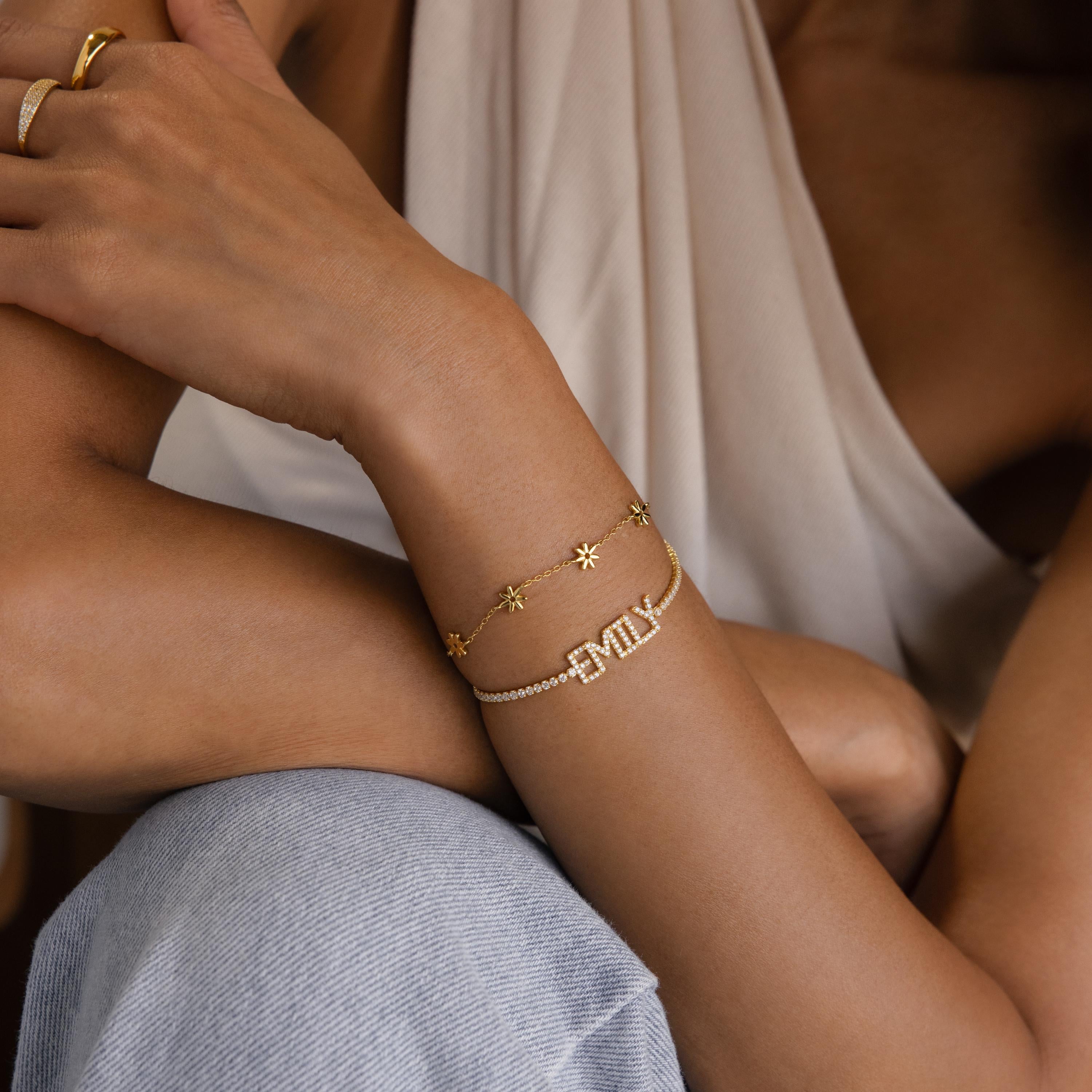 A woman sits with her arms crossed, wearing gold bracelets—including a personalized bracelet with “EMILY” and the Pave Name Tennis Bracelet.