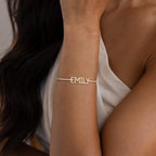 A woman wears the Pave Name Tennis Bracelet, a gold chain personalized with "EMILY" in bold capital letters.