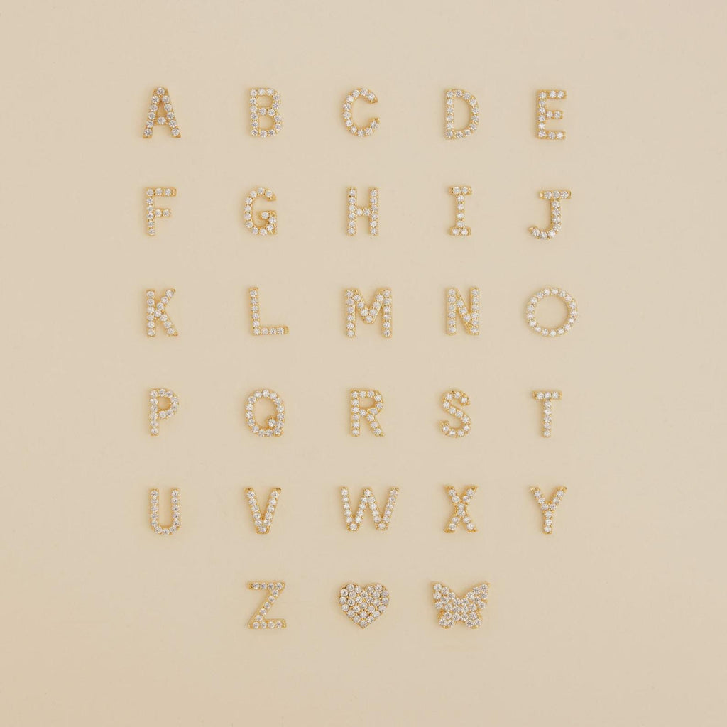 Gold and pearl alphabet letters, a heart, and a butterfly arranged on beige—perfect inspiration for designing your own Diamond Tennis Initial Bracelet or stacking it with others.