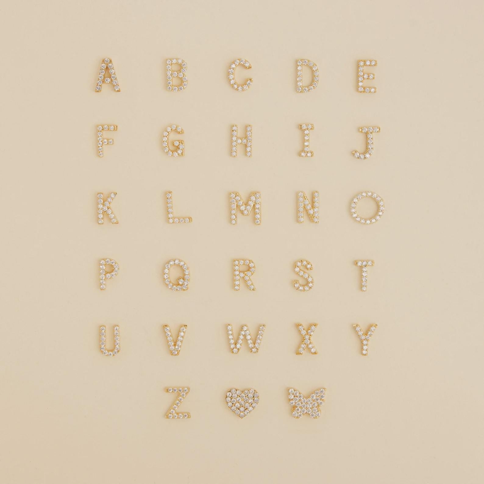 Gold and pearl alphabet letters, a heart, and a butterfly arranged on beige—perfect inspiration for designing your own Diamond Tennis Initial Bracelet or stacking it with others.