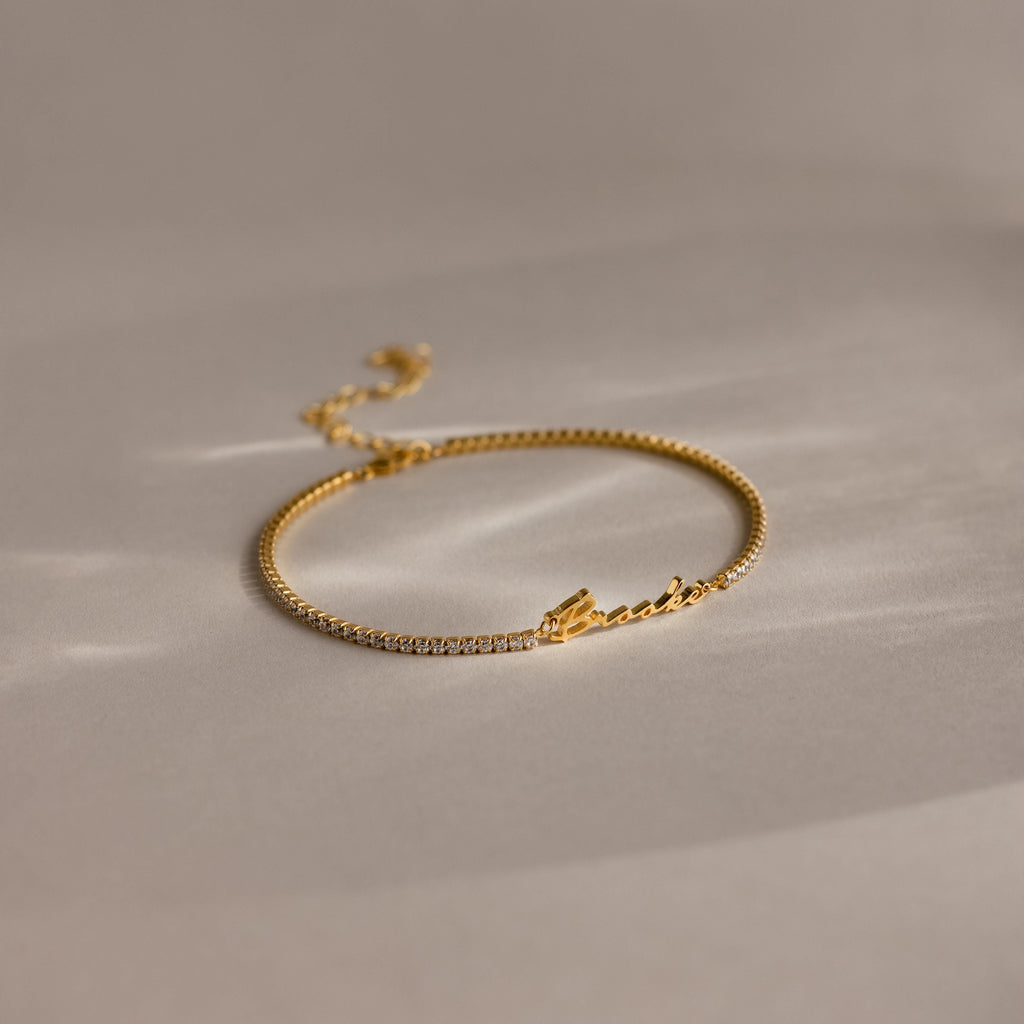 The Venice Name Tennis Bracelet features delicate gold, small diamonds, and a personalized script name detail, elegantly displayed on a neutral background.