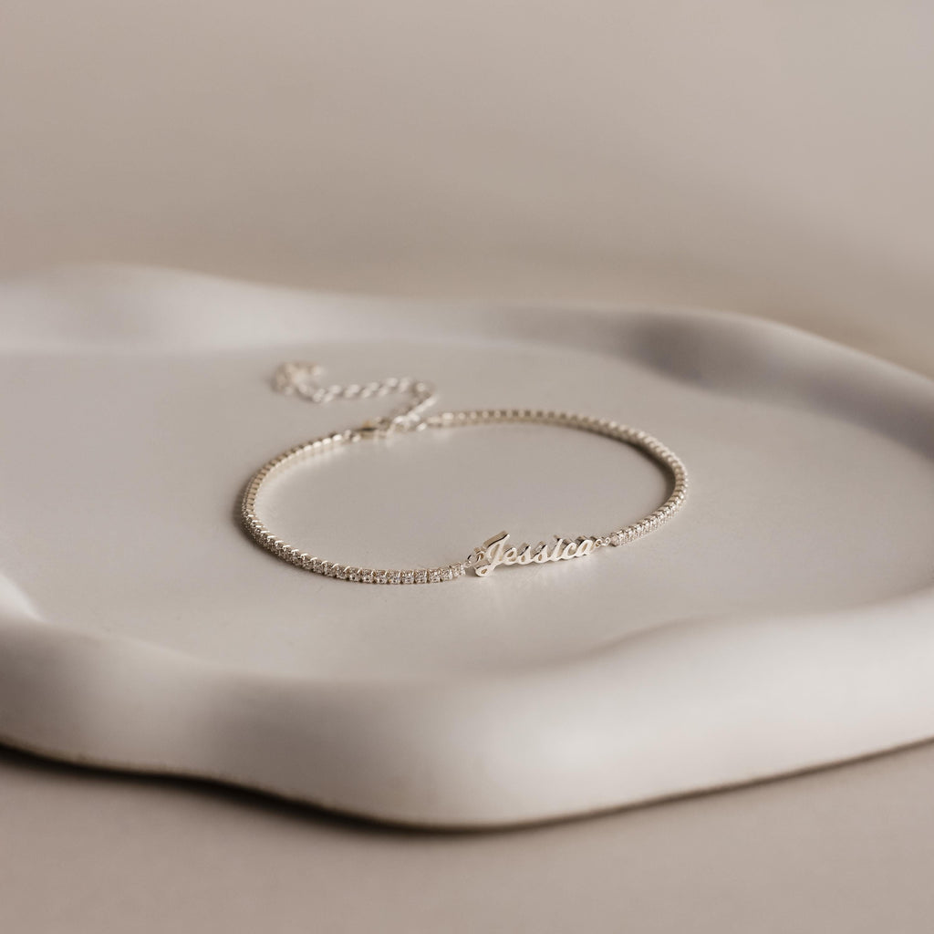 A Venice Name Tennis Bracelet in silver, personalized with "Jessica" as the nameplate charm, is displayed elegantly on a white wavy-edged dish.