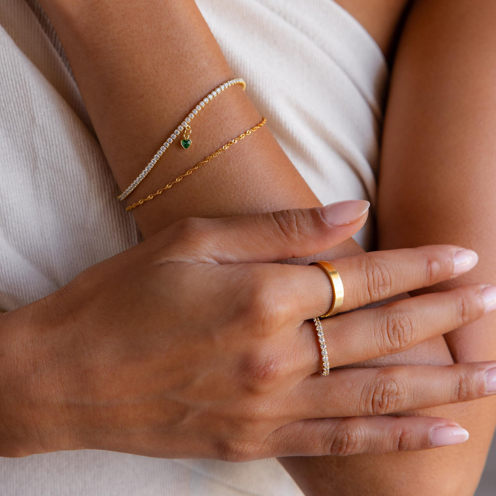 Close-up of hands wearing gold and diamond bracelets—featuring the Heart Birthstone Tennis Bracelet—and personalized charm rings, resting on light fabric.